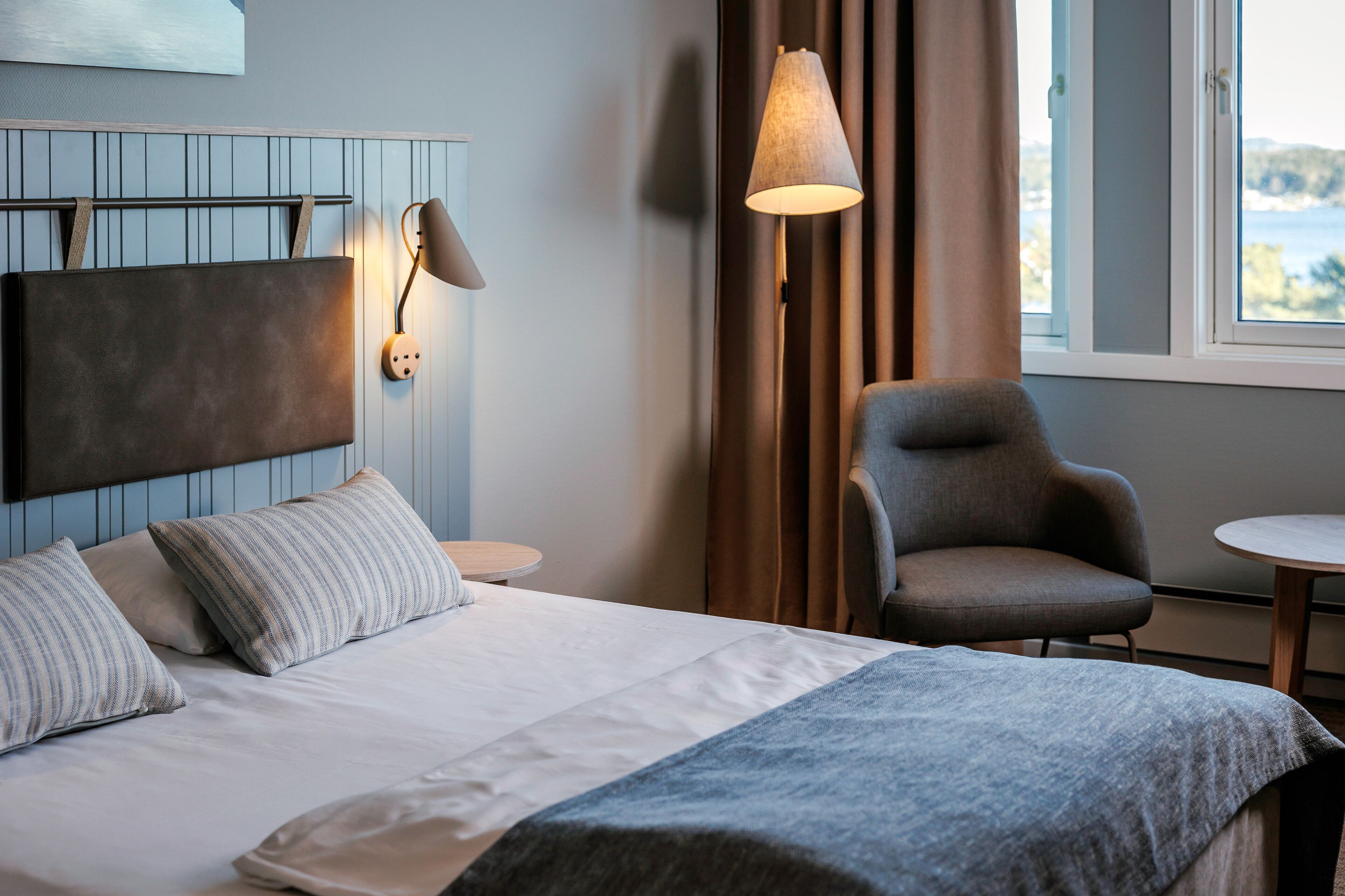 A hotel room with a bed, a chair, a lamp, and a view of the sea through the window.
