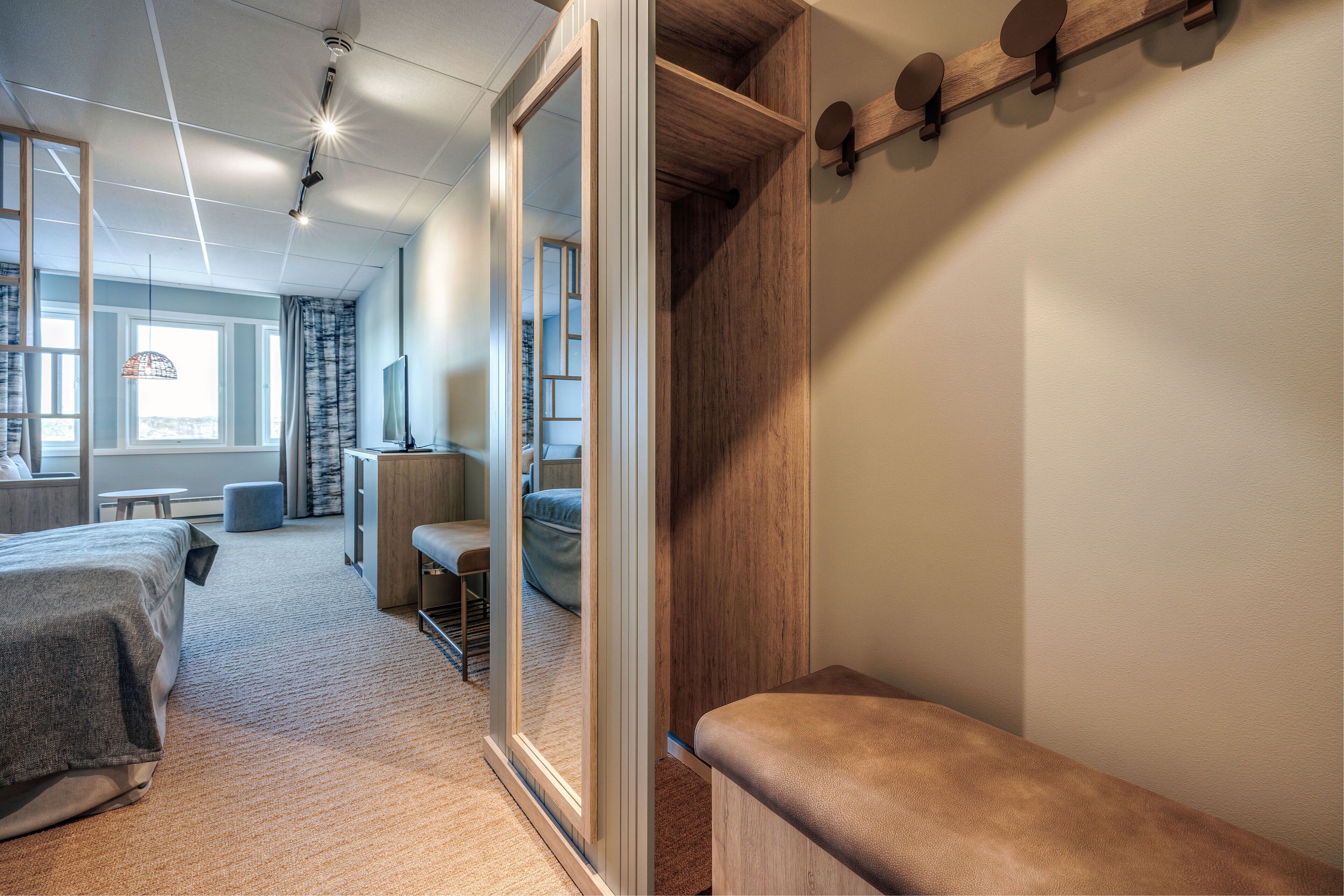 Une chambre d’hôtel moderne, dotée d’un grand lit, d’un bureau, d’une chaise et d’un banc. Une fenêtre offre une vue sur la ville, et une étagère en bois est agrémentée d’objets décoratifs.
