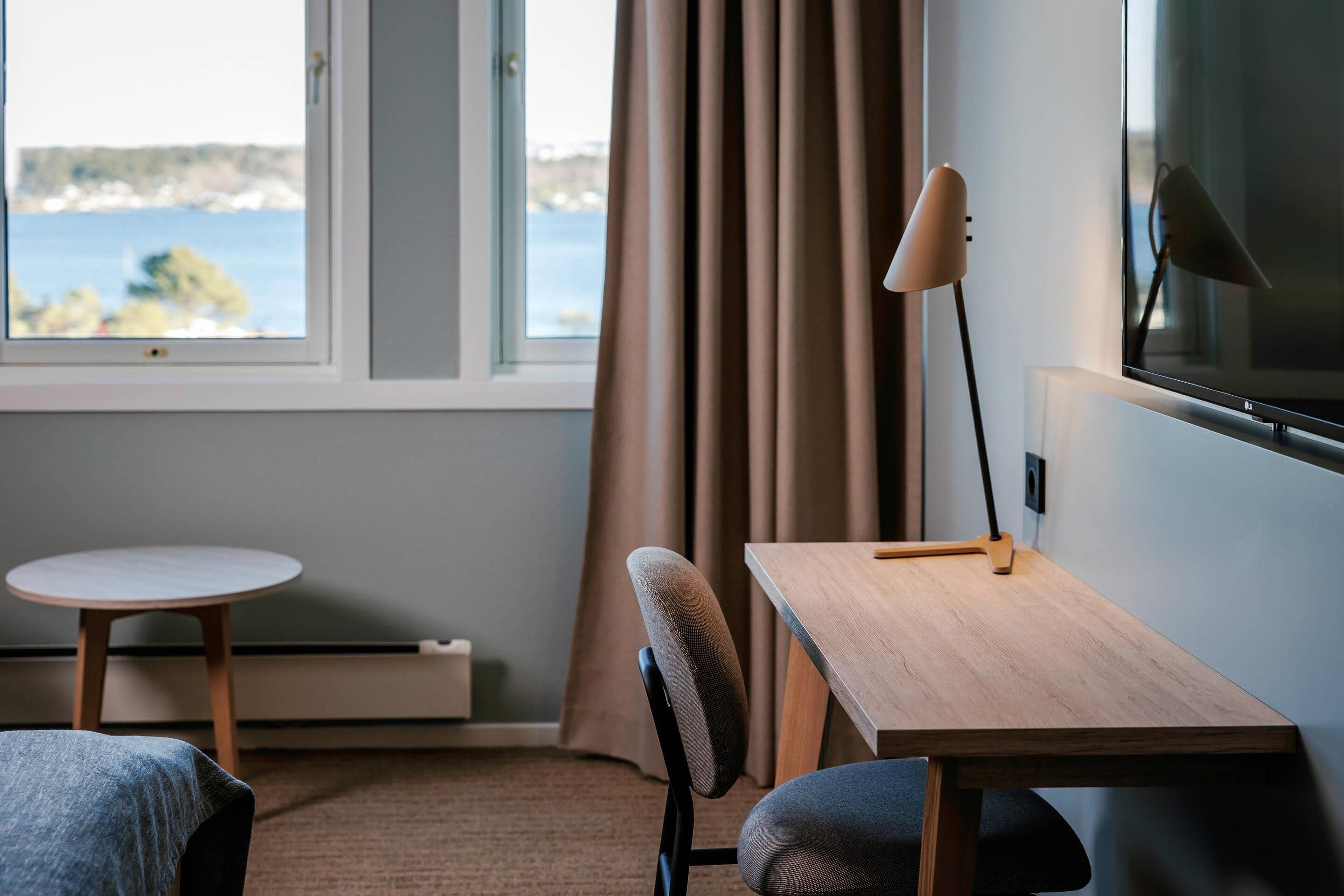 A room with a bed, a desk, a chair, a lamp, a TV, a window with curtains, and a view of the sea.