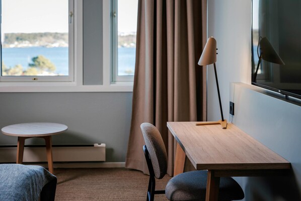 Una habitación con cama, escritorio, silla, lámpara, televisor y ventana con cortinas, con vista al mar.