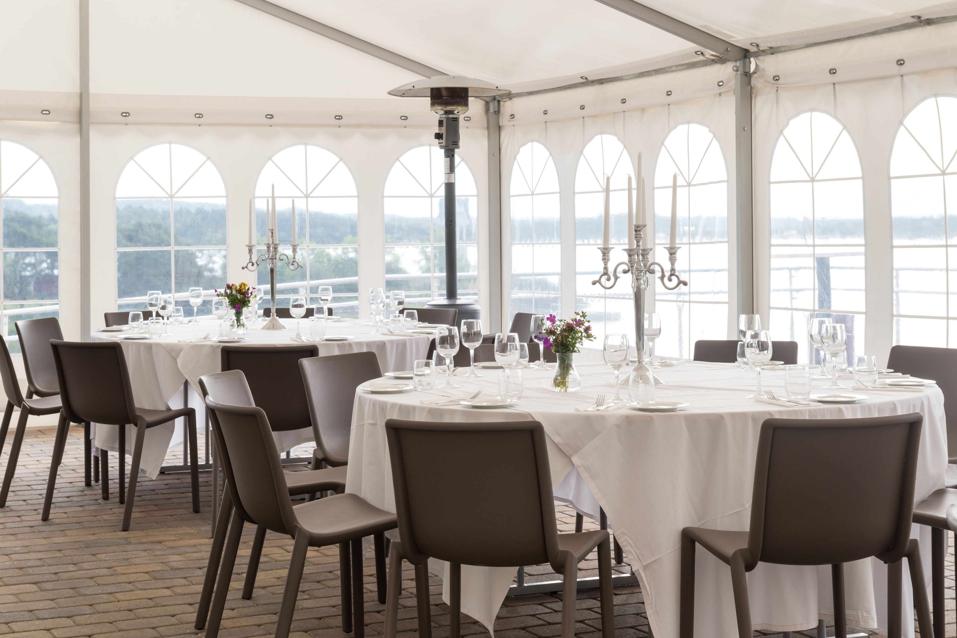 Un área de comedor con carpas, mesas redondas preparadas para una comida, ventanas amplias que ofrecen vistas a un cuerpo de agua y un candelabro colgado del techo.