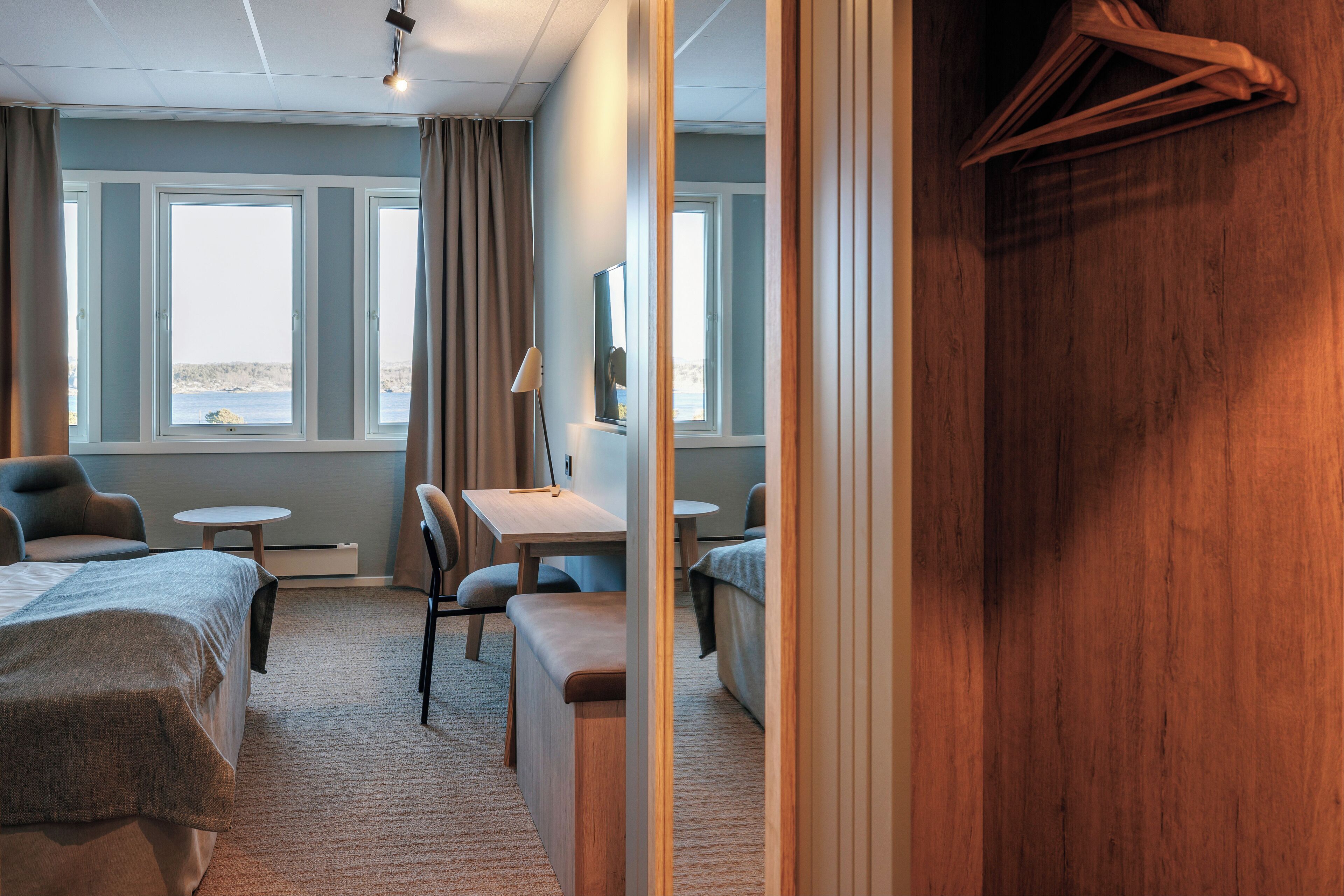 Een hotelkamer met een bed, een bureau, een stoel, een klein tafeltje en uitzicht op zee door grote ramen.