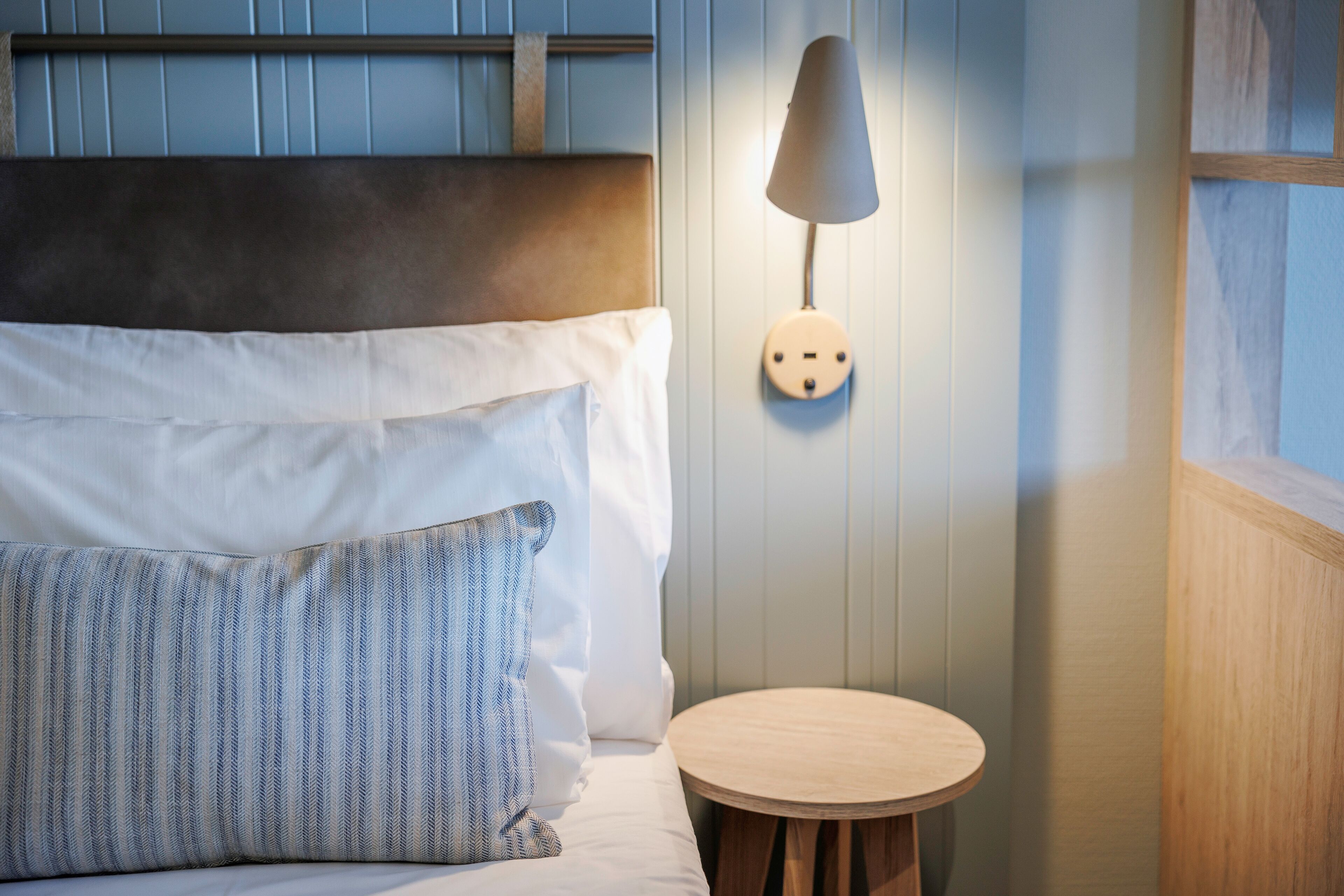 A neatly made bed with white and striped pillows, a round wooden side table, and a wall-mounted lamp.