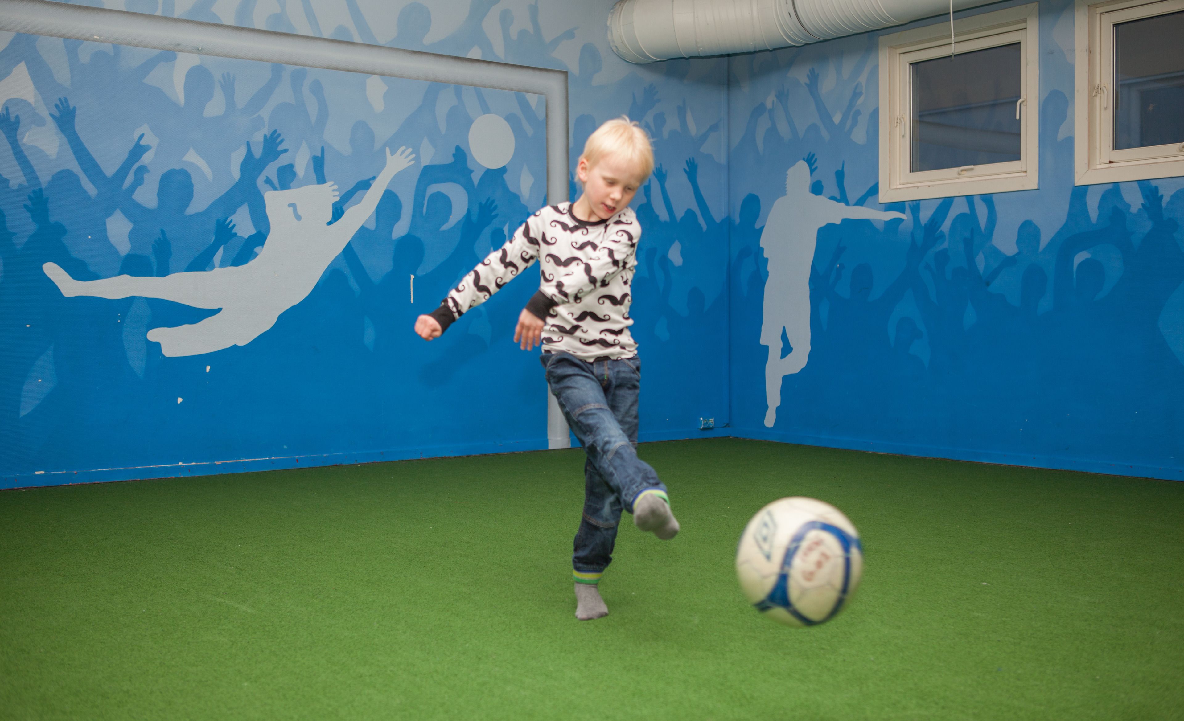 Un niño juega con una pelota de fútbol en una habitación con paredes azules y suelo verde.