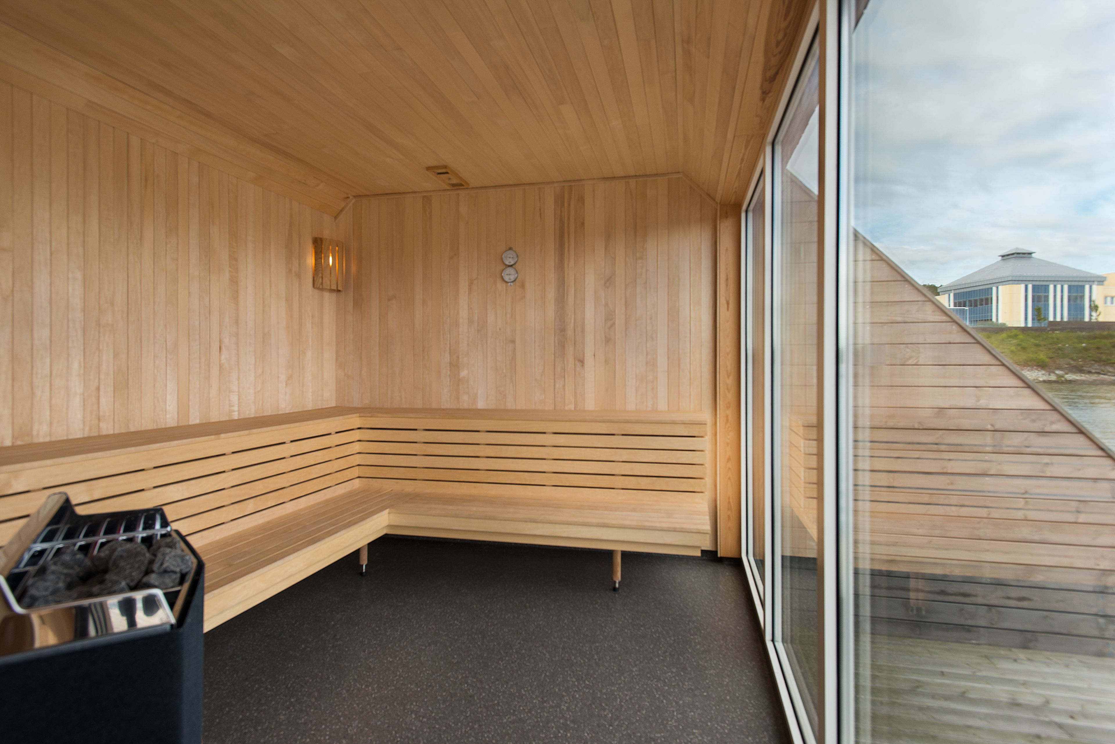 Un sauna aux murs et aux bancs en bois, avec un sol noir et une fenêtre donnant sur les bâtiments environnants.