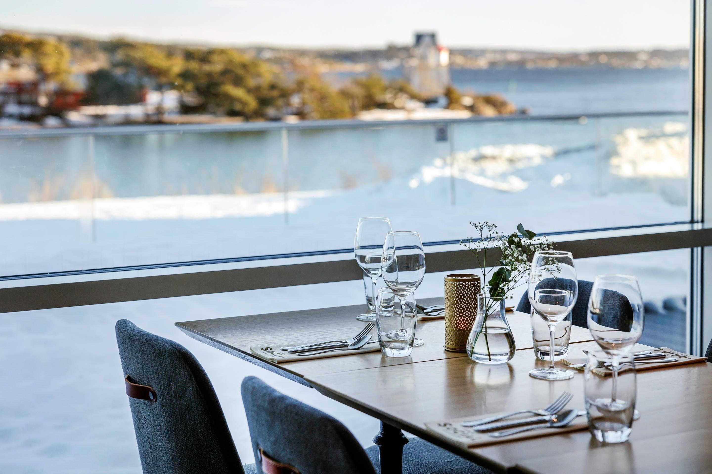 Una mesa de comedor con sillas dispuesta para dos personas, con vista a un cuerpo de agua donde se ven barcos y edificios a lo lejos.
