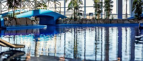 En inomhuspool med en blå vattenrutschkana, palmer och utsikt över omgivningen genom stora fönster.
