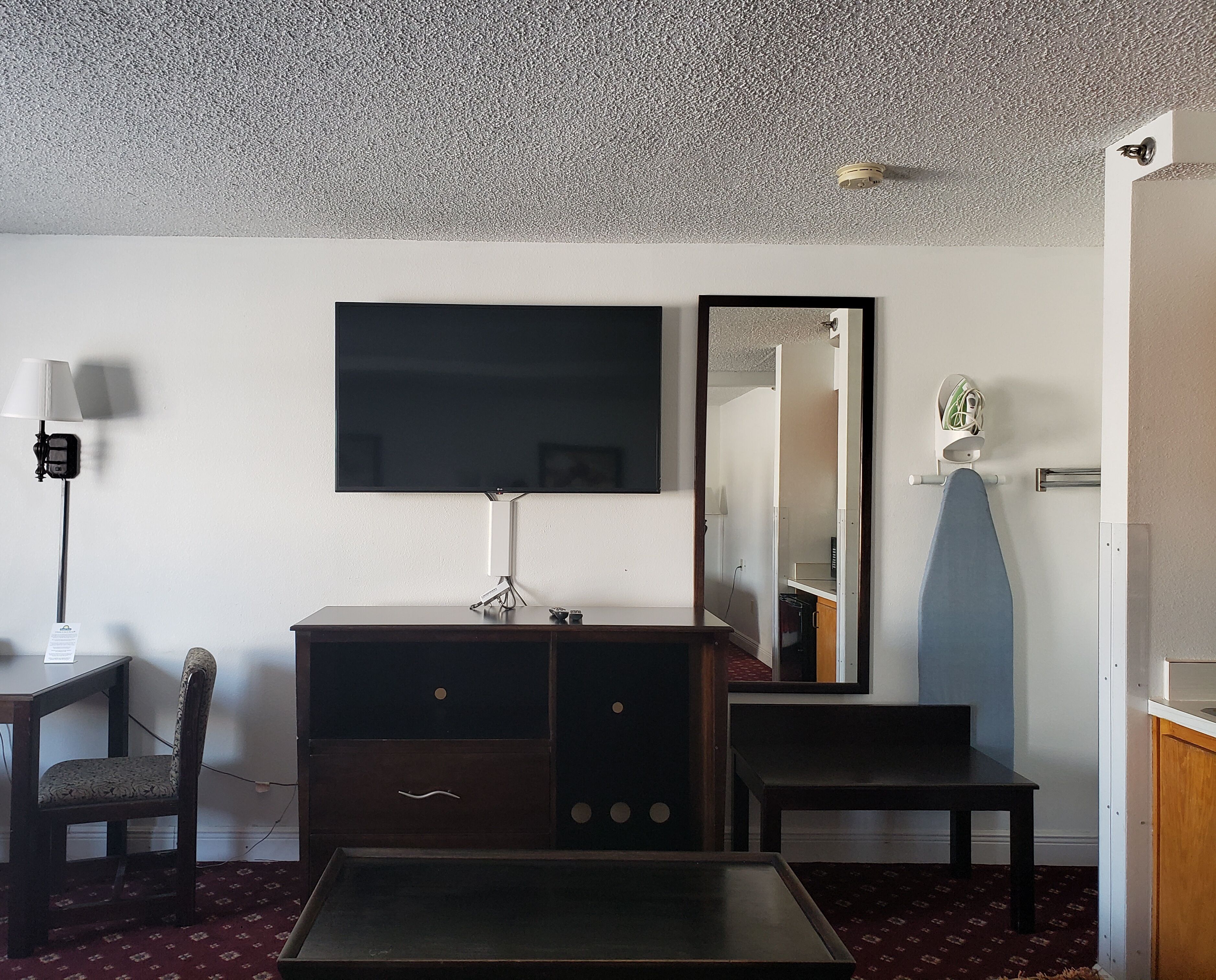 Une chambre d’hôtel dotée d’un grand téléviseur à écran plat fixé au mur, d’une commode en bois, d’une petite table, d’une chaise et d’un miroir qui reflète la pièce.