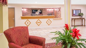 A reception area with a wooden counter, a red patterned armchair, and a potted plant with red flowers. - Days Inn by Wyndham Lake Village (Lake Village)