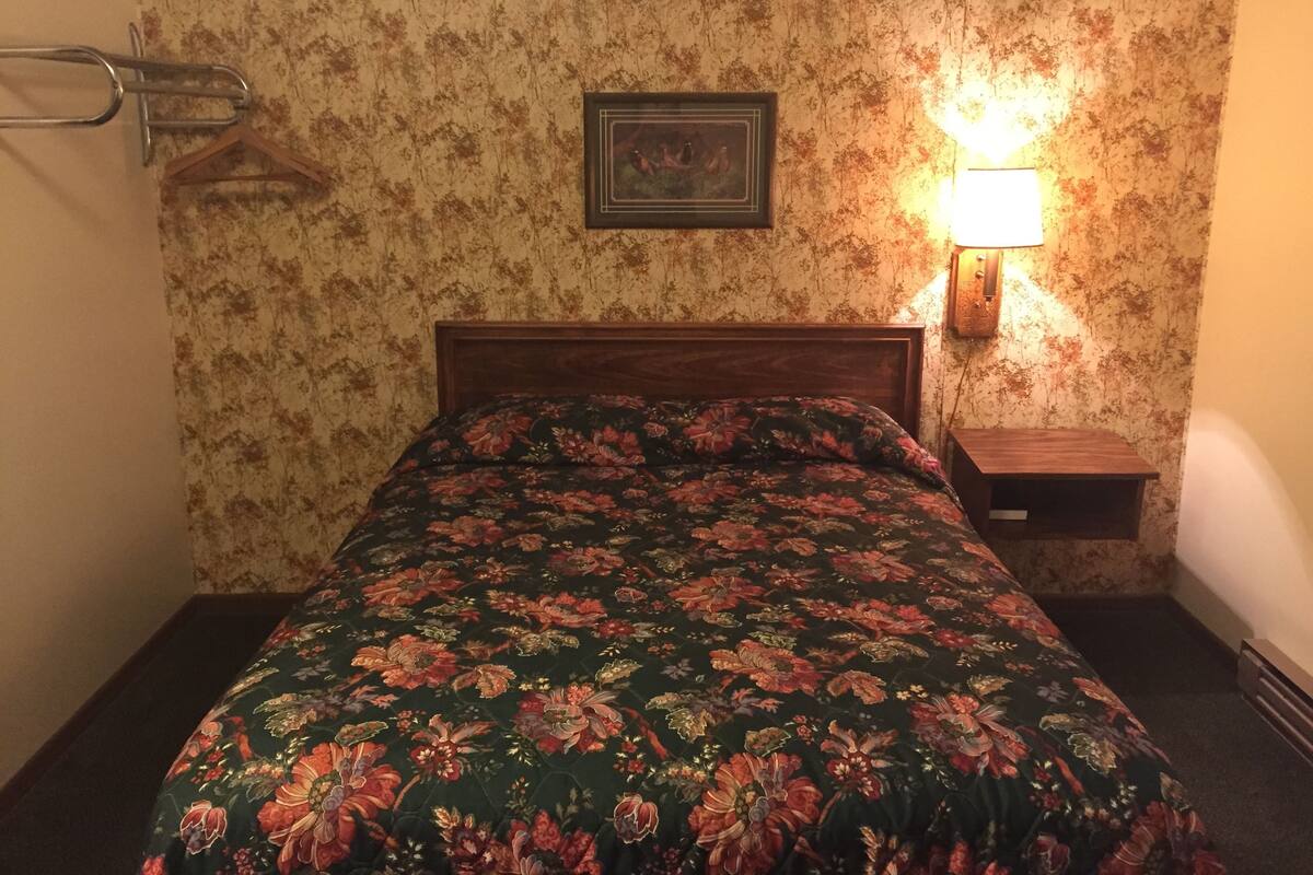 Quarto com edredom floral, cabeceira de madeira, mesa de cabeceira com abajur e um quadro na parede.