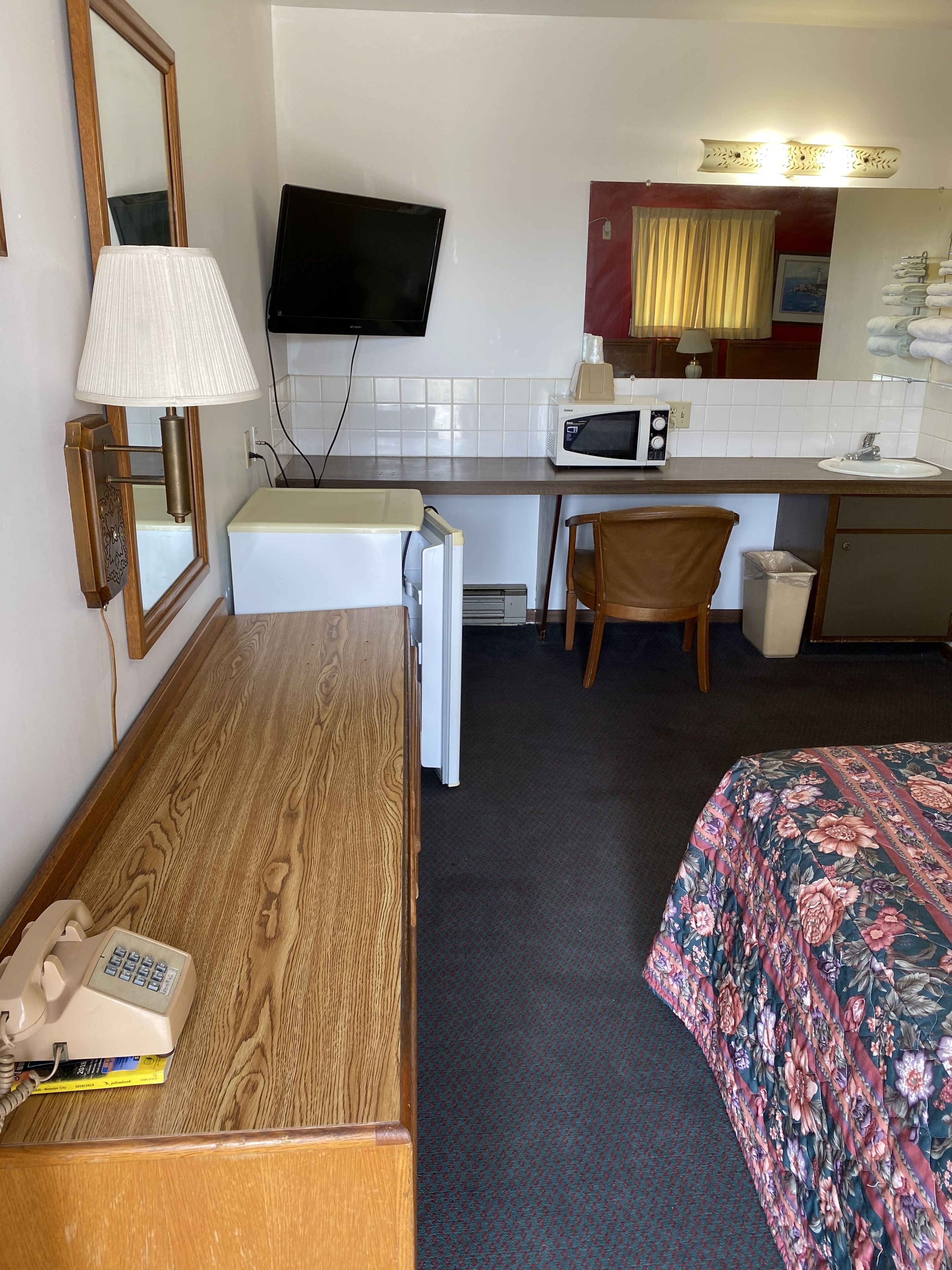 A compact hotel room with a wooden desk, a bed with floral bedding, a television, a microwave, and a small bathroom area.