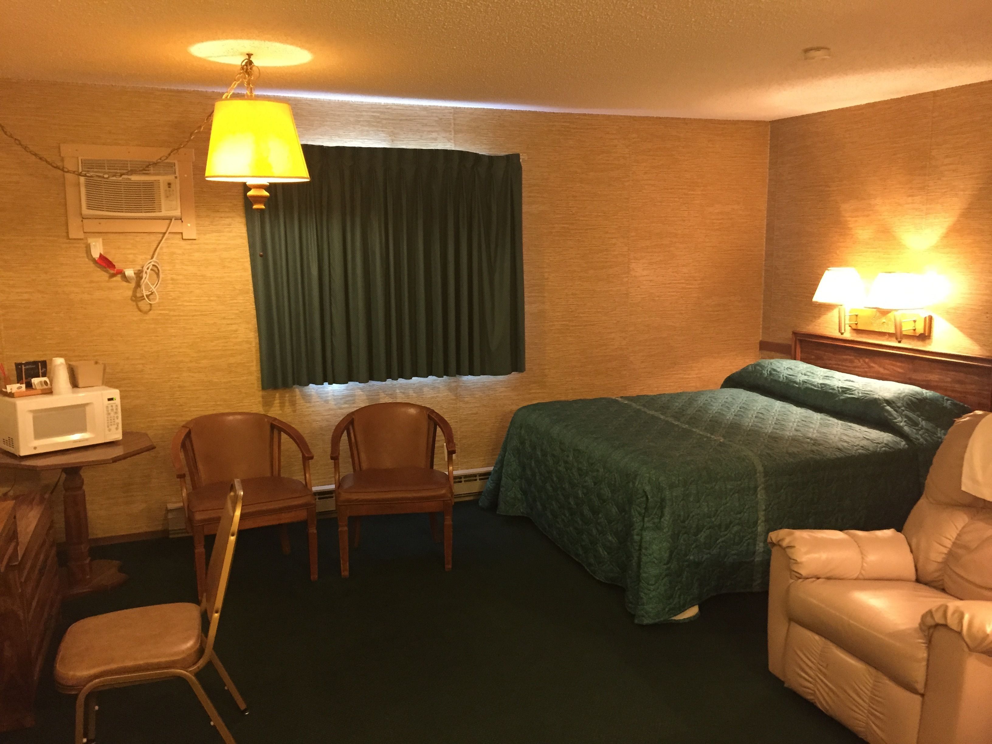 Une chambre d’hôtel avec un lit, deux chaises, un bureau avec un four à micro-ondes, une lampe et une fenêtre avec des rideaux.