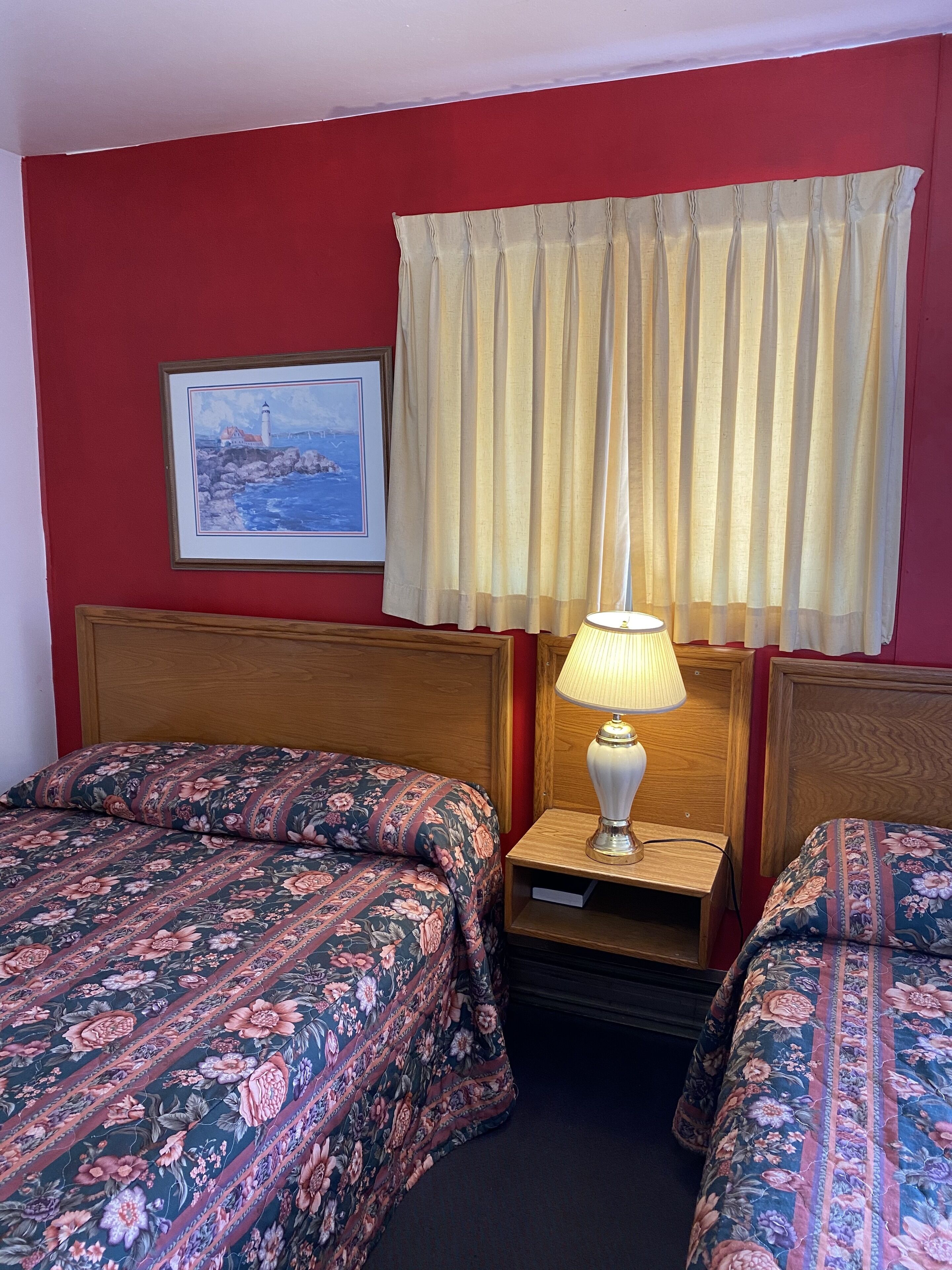 A hotel room with two beds, a nightstand with a lamp, a framed painting on the wall, and a window with curtains.