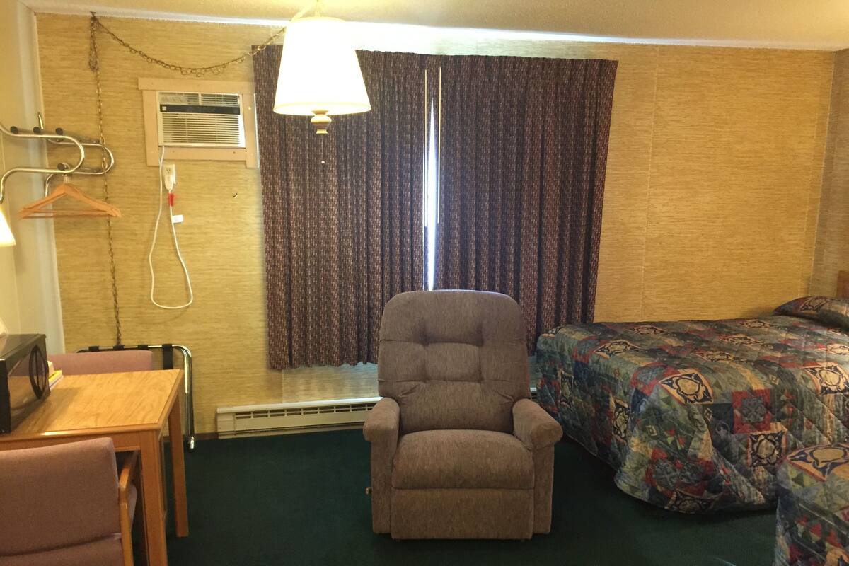 Quarto de hotel com uma cama de solteiro, uma cadeira, uma escrivaninha e um candeeiro suspenso.