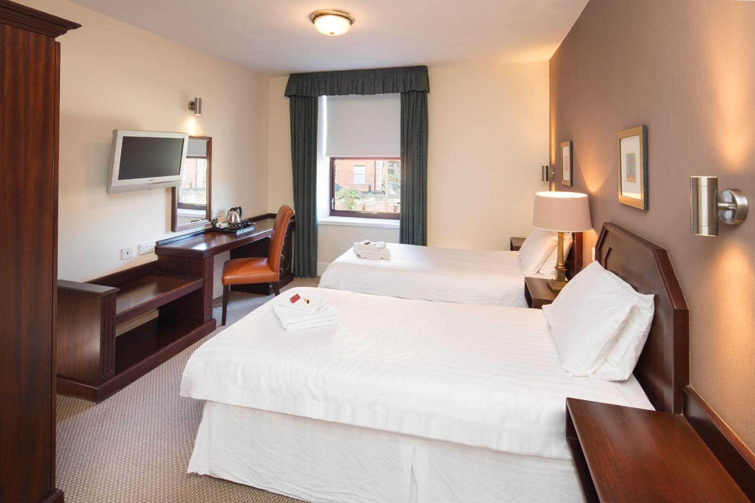 A hotel room with two beds, a desk, a chair, a TV, and a window with curtains.