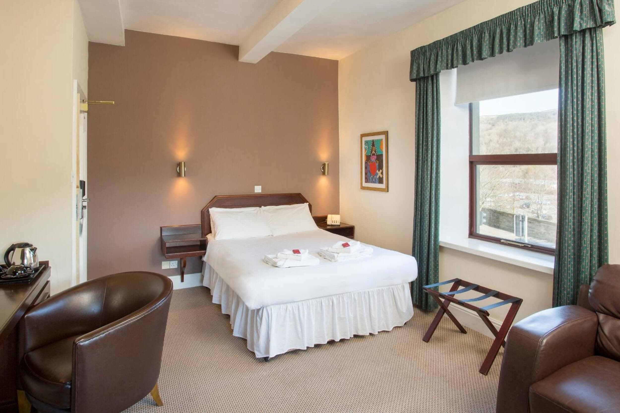 A hotel room with a large bed, a desk, a chair, a nightstand, a window with curtains, and a painting on the wall.
