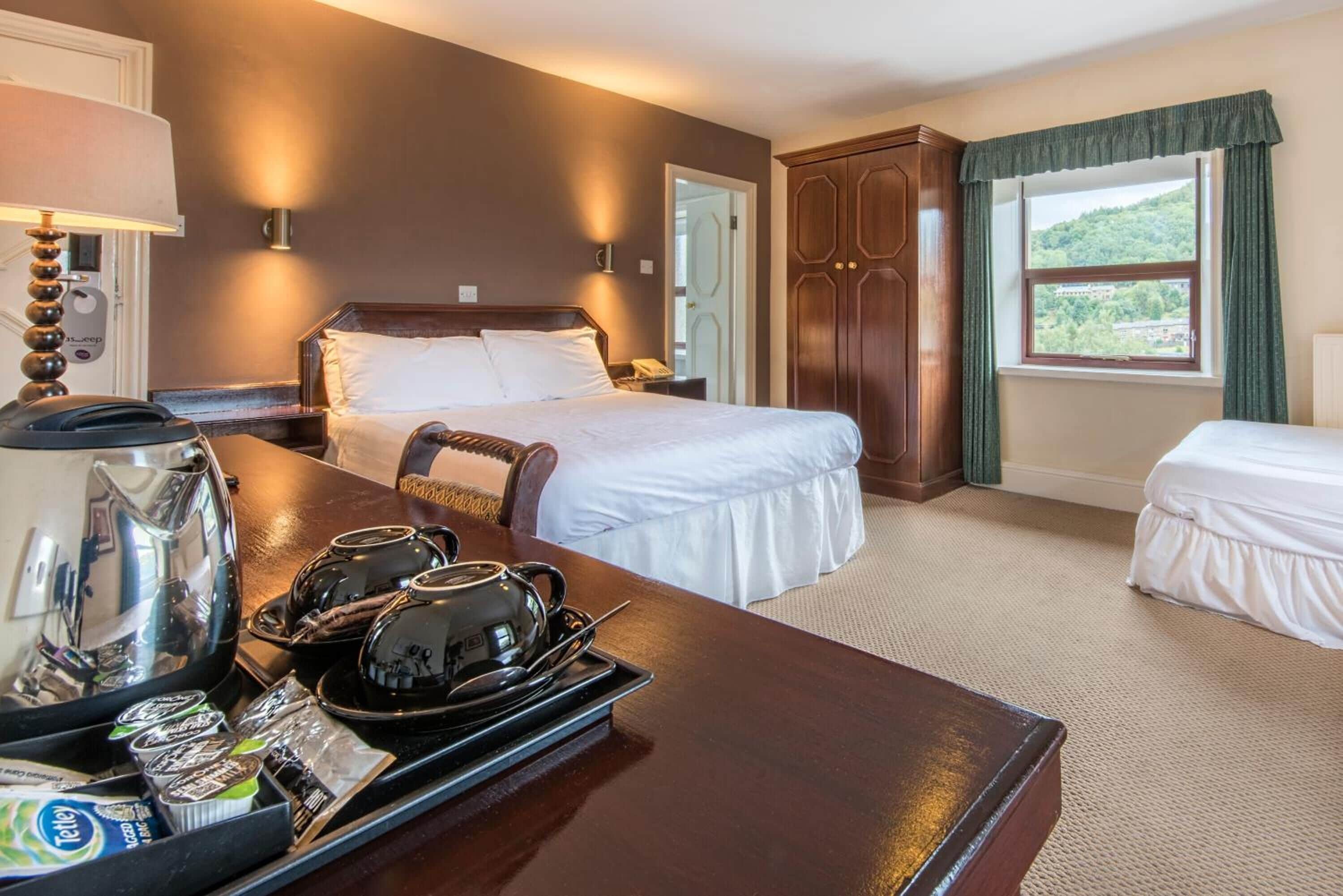 A hotel room with a large bed, a bedside table with a kettle and tea packets, a wooden wardrobe, and a window with a view of greenery.