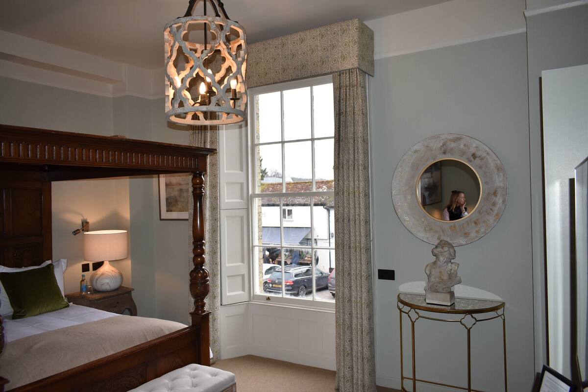 A bedroom with a four-poster bed, a bedside table with a lamp, a window with curtains, and a round mirror on a small table.