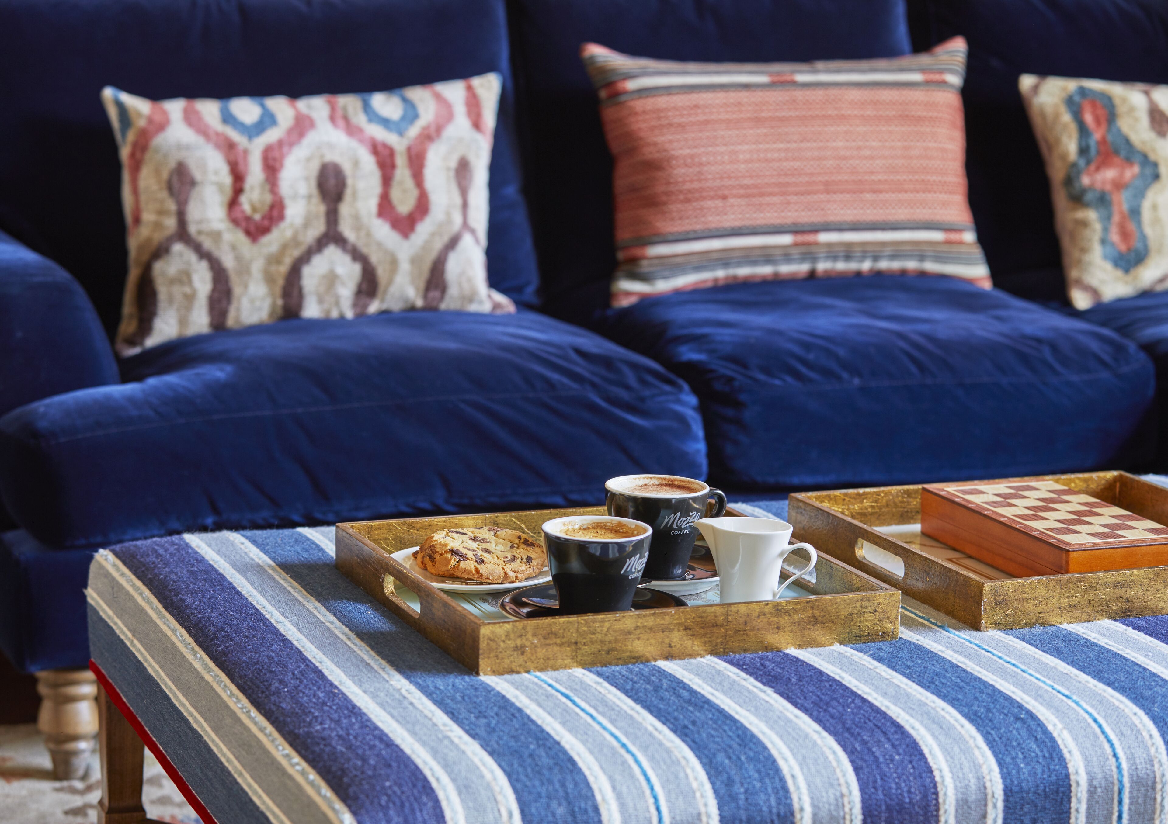 A cozy seating area with a blue sofa and ottoman, a tray with a muffin, coffee cups, and a chessboard.