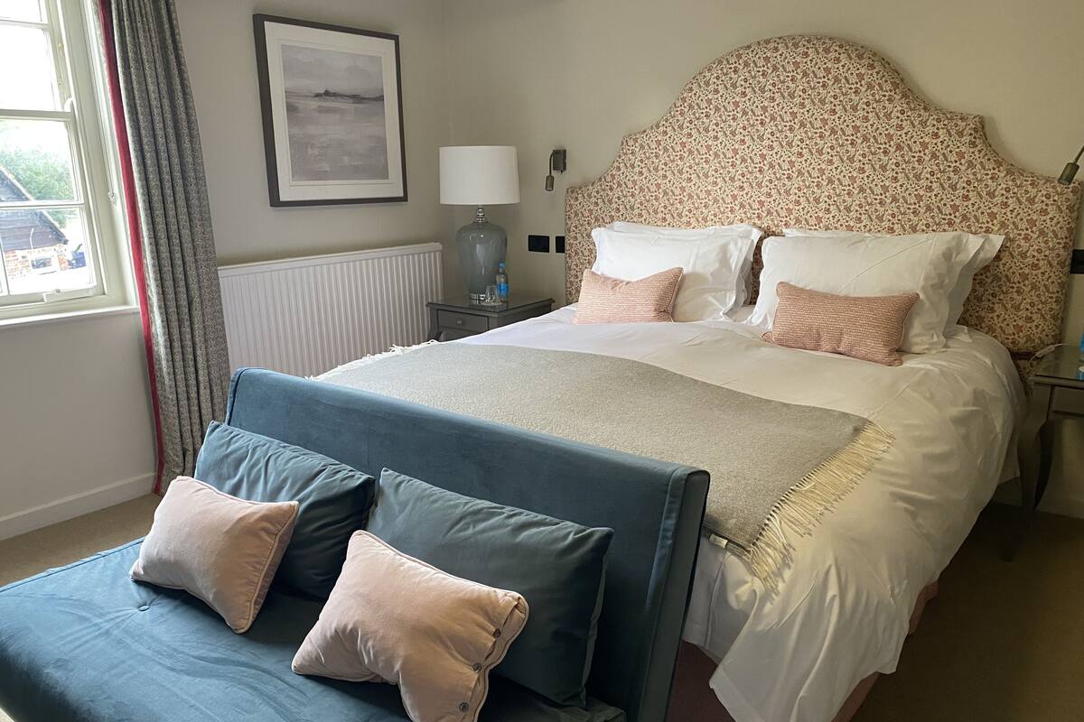 A hotel room with a large bed, a bedside table with a lamp, a framed picture on the wall, and a window with curtains.