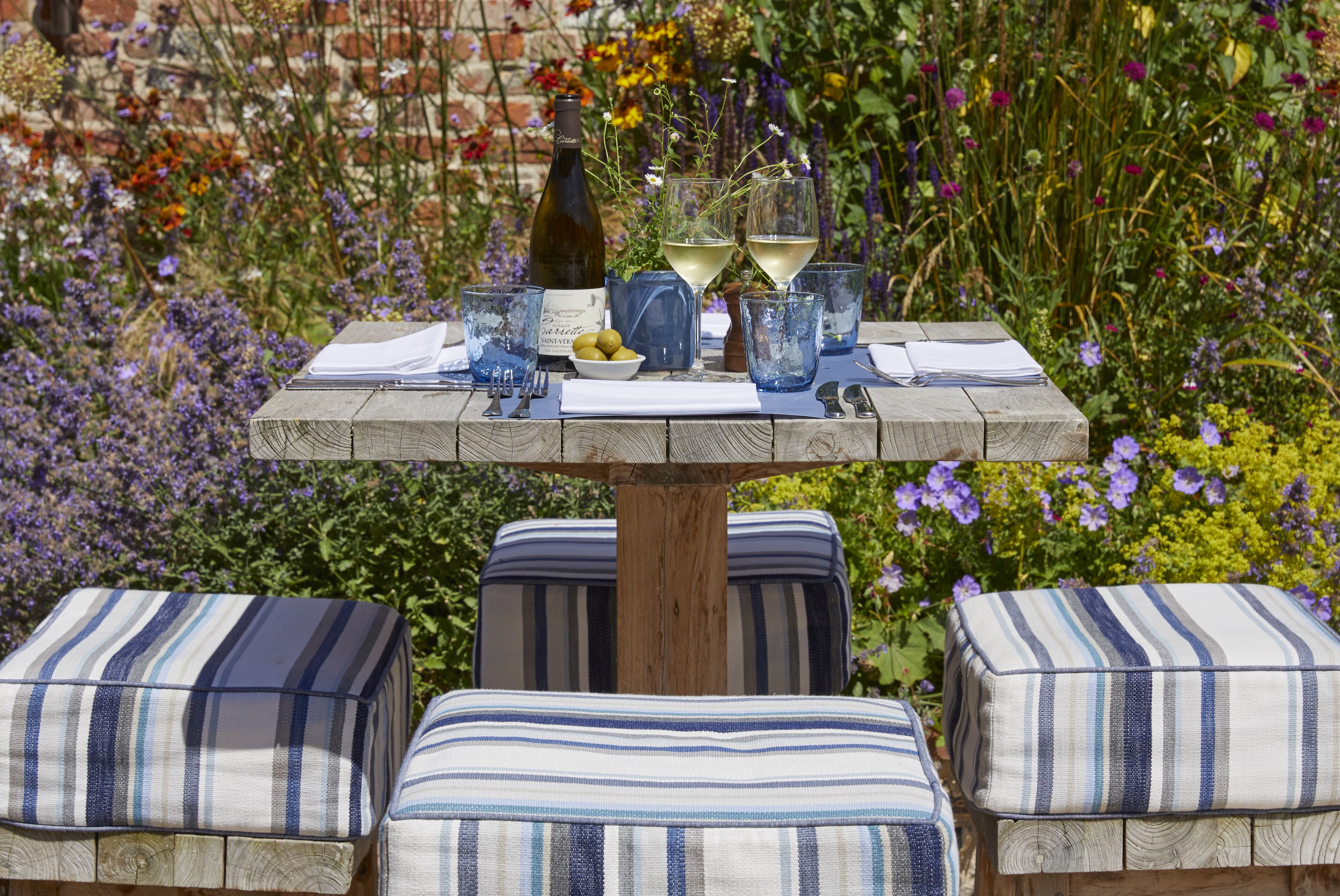A wooden table with a bottle of wine, glasses, and a bowl of olives. There are striped cushions on the table.