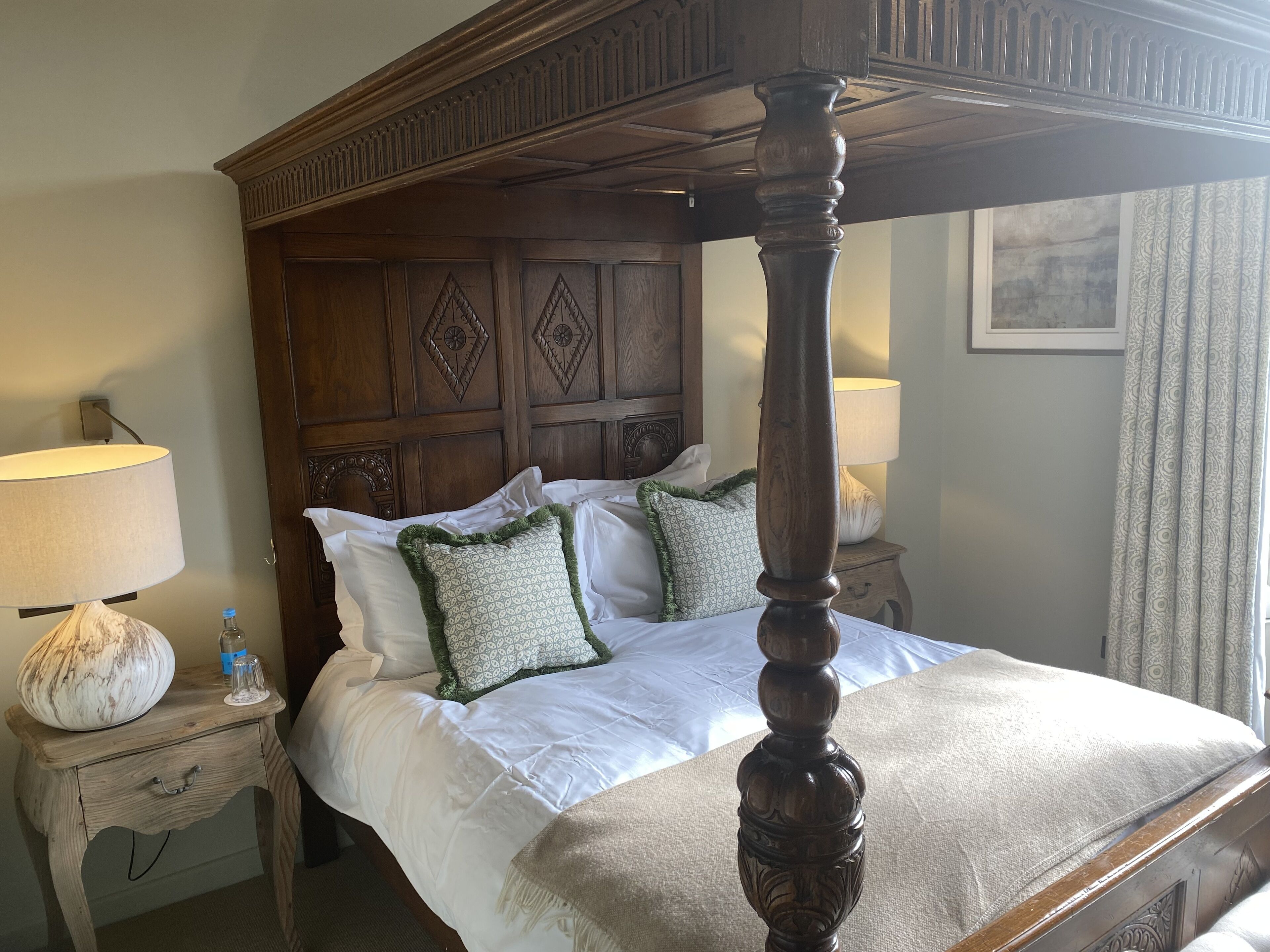 A four-poster bed with a wooden headboard, white bedding, and decorative pillows. There are two bedside tables with lamps and a decorative vase.
