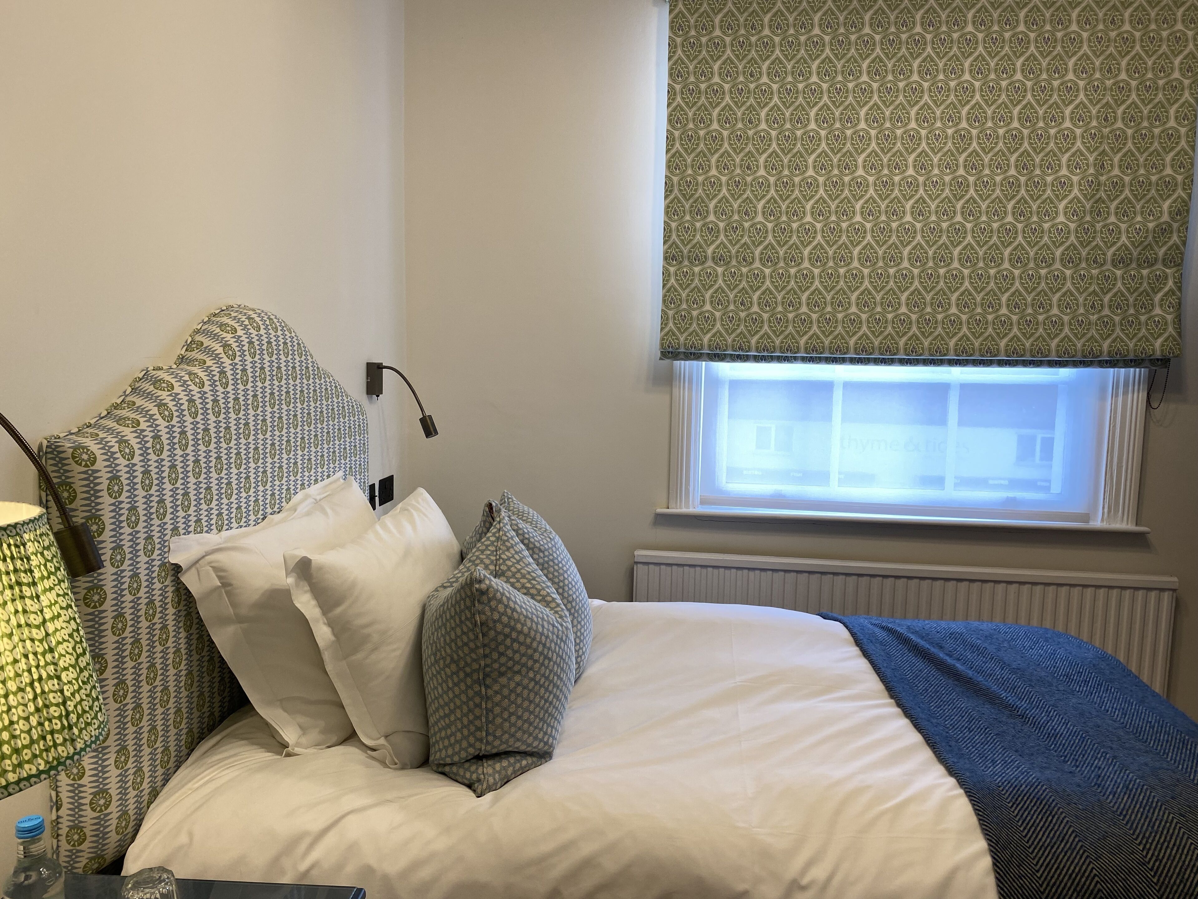 A neatly made bed with patterned pillows and a blue throw blanket, a bedside lamp, and a window with a patterned blind.