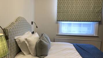 A neatly made bed with patterned pillows and a blue throw blanket, a bedside lamp, and a window with a patterned blind.