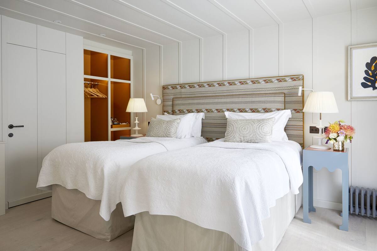 A double bed with white linens, a bedside table with a lamp, a small side table with a vase of flowers, and a wardrobe with a mirror.