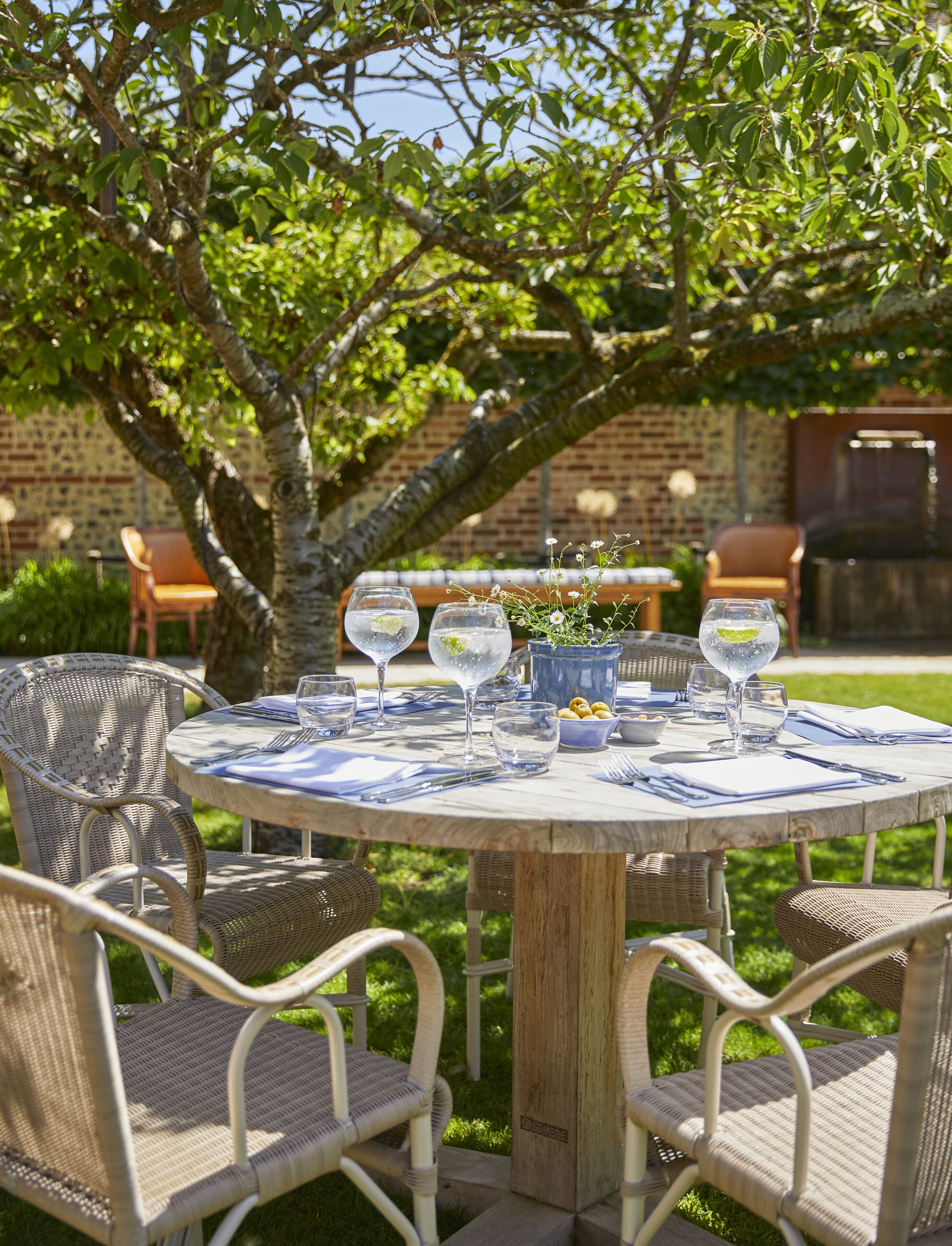 A round wooden table set outdoors with wicker chairs, surrounded by greenery and a brick wall.