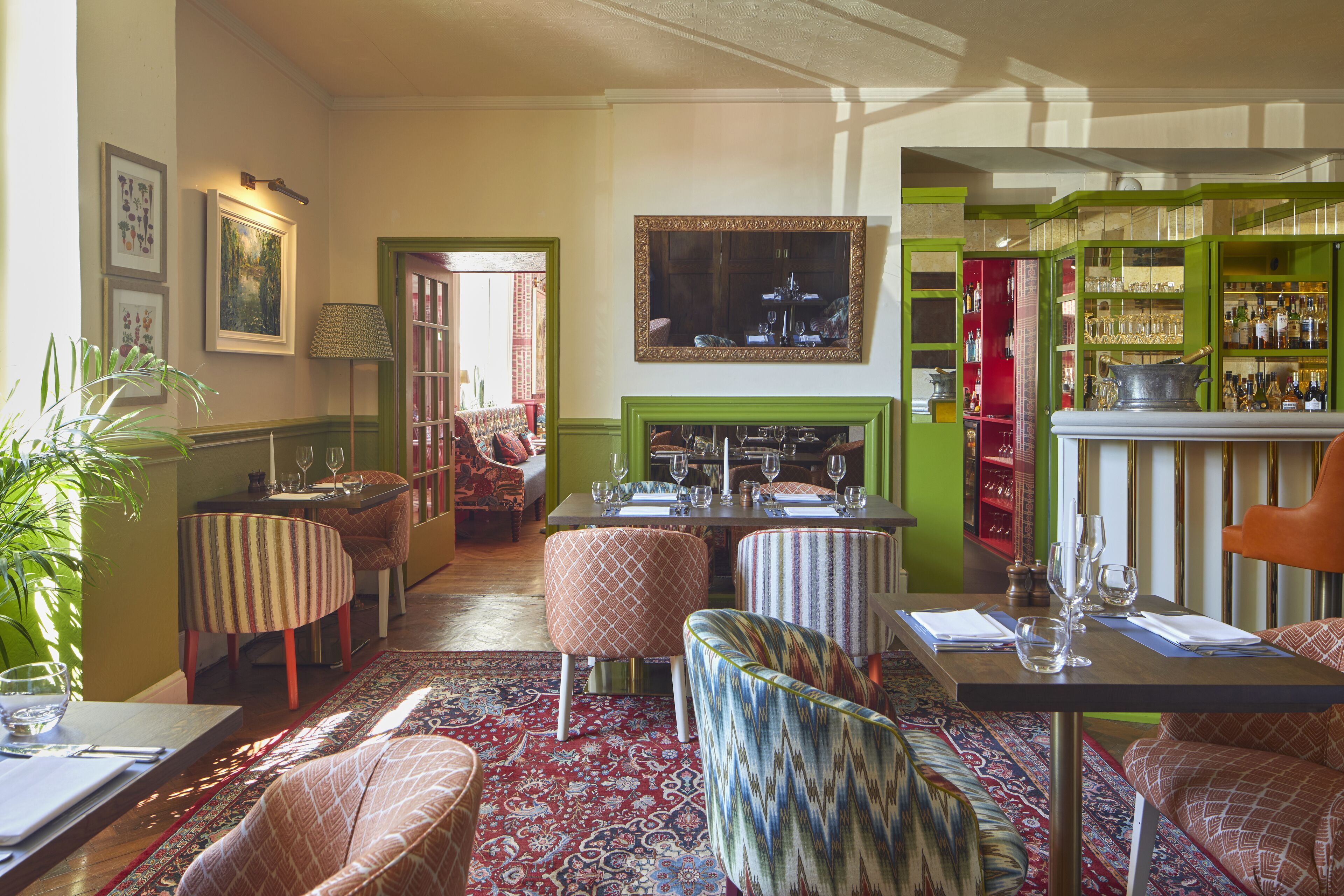 A dining area with a table set for two, a patterned rug, and a green-framed mirror above the fireplace.