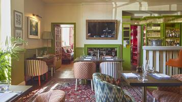 A dining area with a table set for two, a patterned rug, and a green-framed mirror above the fireplace.