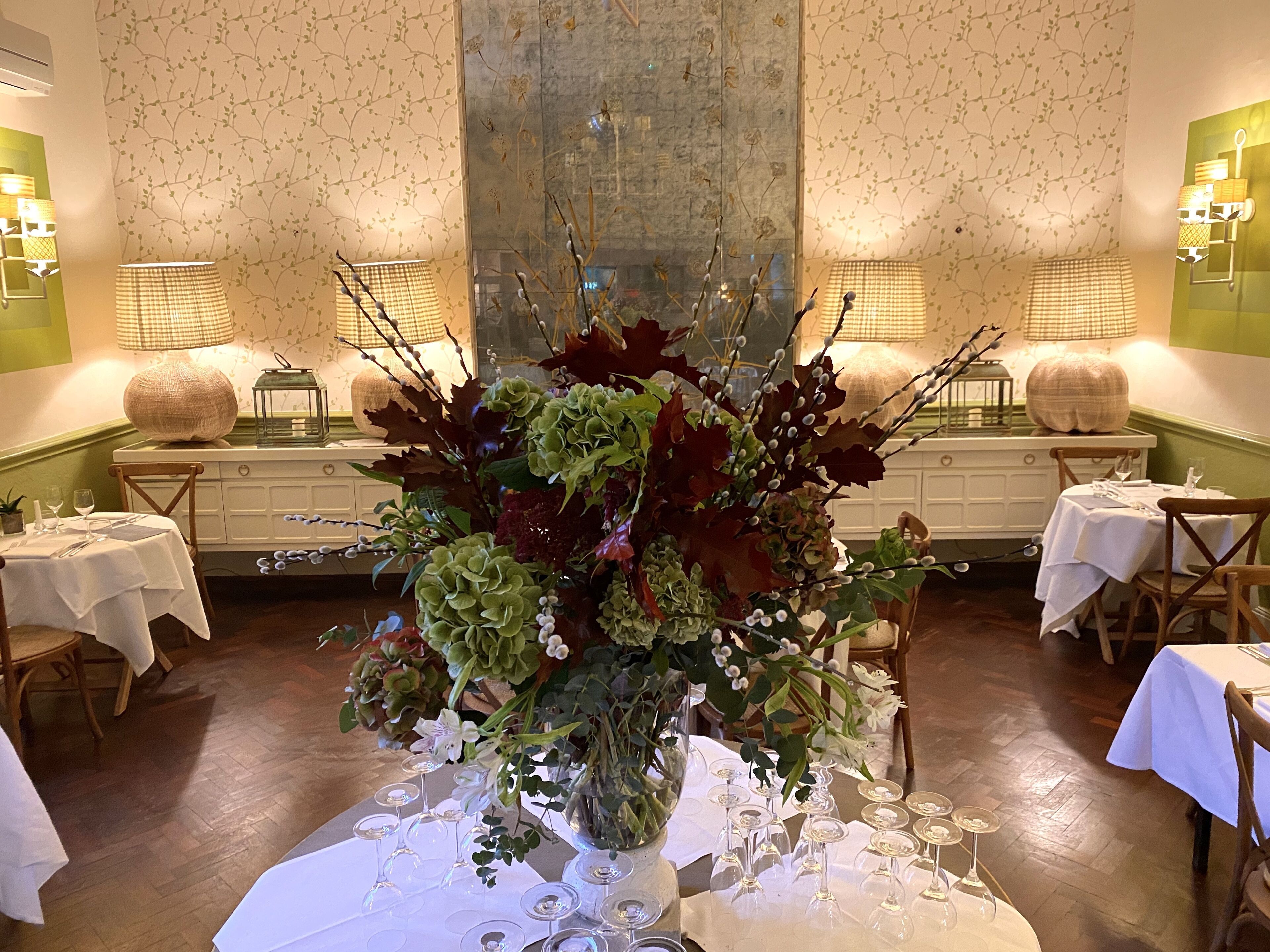 Un comedor con mesas dispuestas para una comida, un gran centro de mesa floral y lámparas fijadas a la pared.