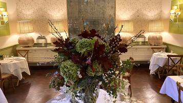 Un comedor con mesas dispuestas para una comida, un gran centro de mesa floral y lámparas fijadas a la pared.