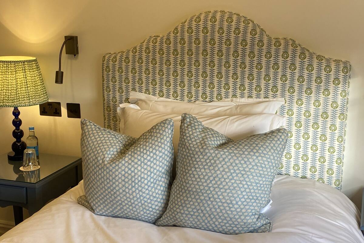 A hotel room with a patterned headboard, a bedside table with a lamp, and two patterned pillows.