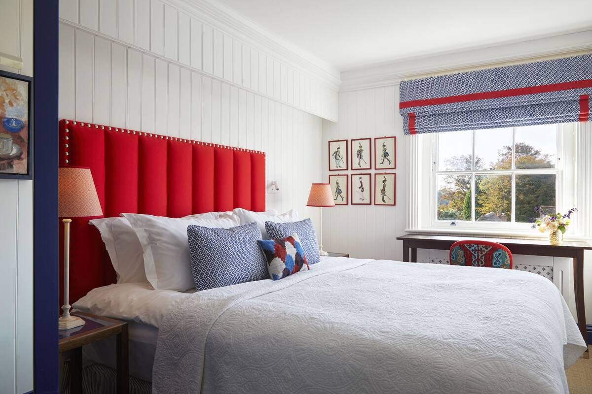 A neatly made bed with a red headboard, white bedding, and a window with a view of trees.