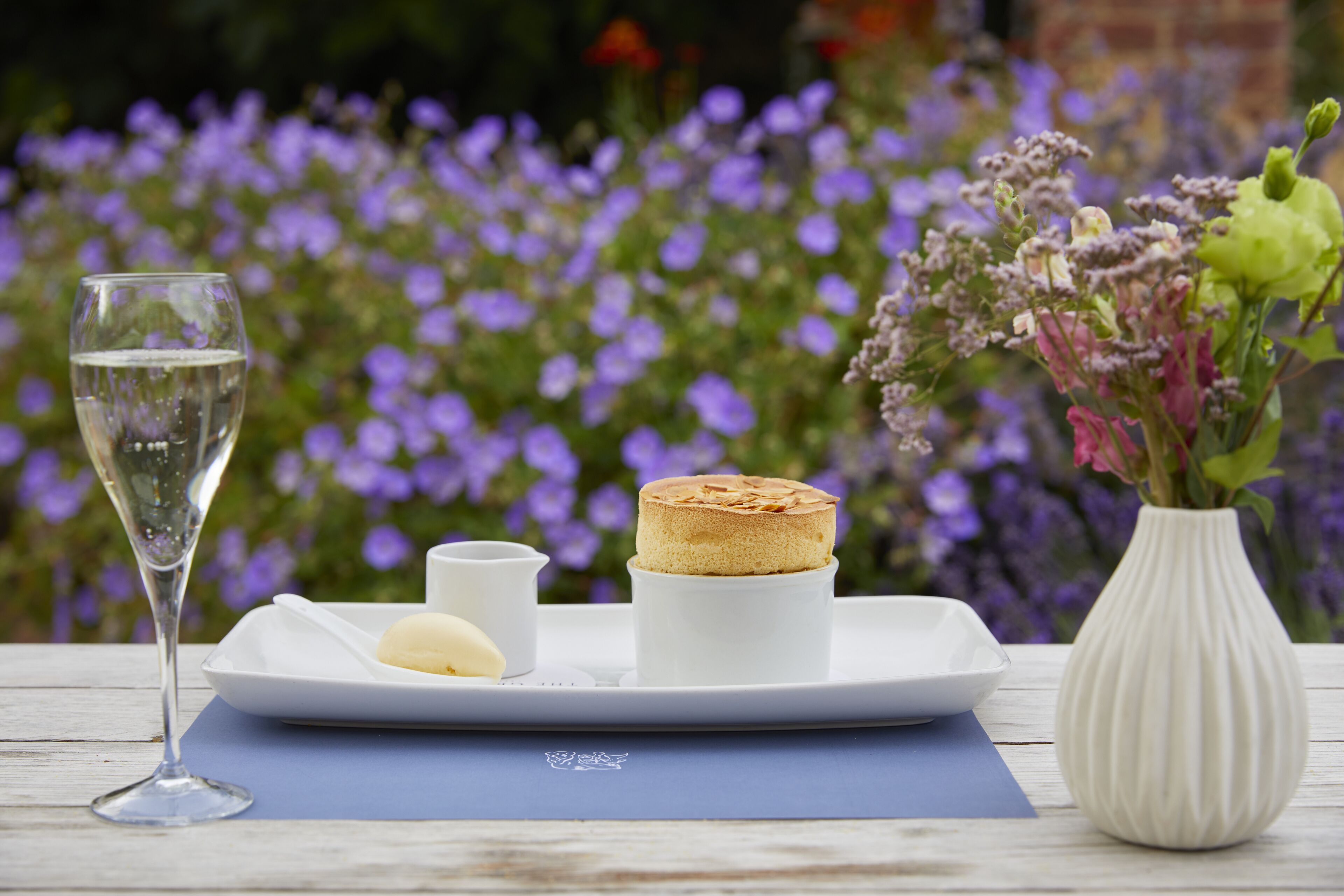 A table setting with a glass of water, a small white dish, a white plate with a cake, and a vase with flowers.