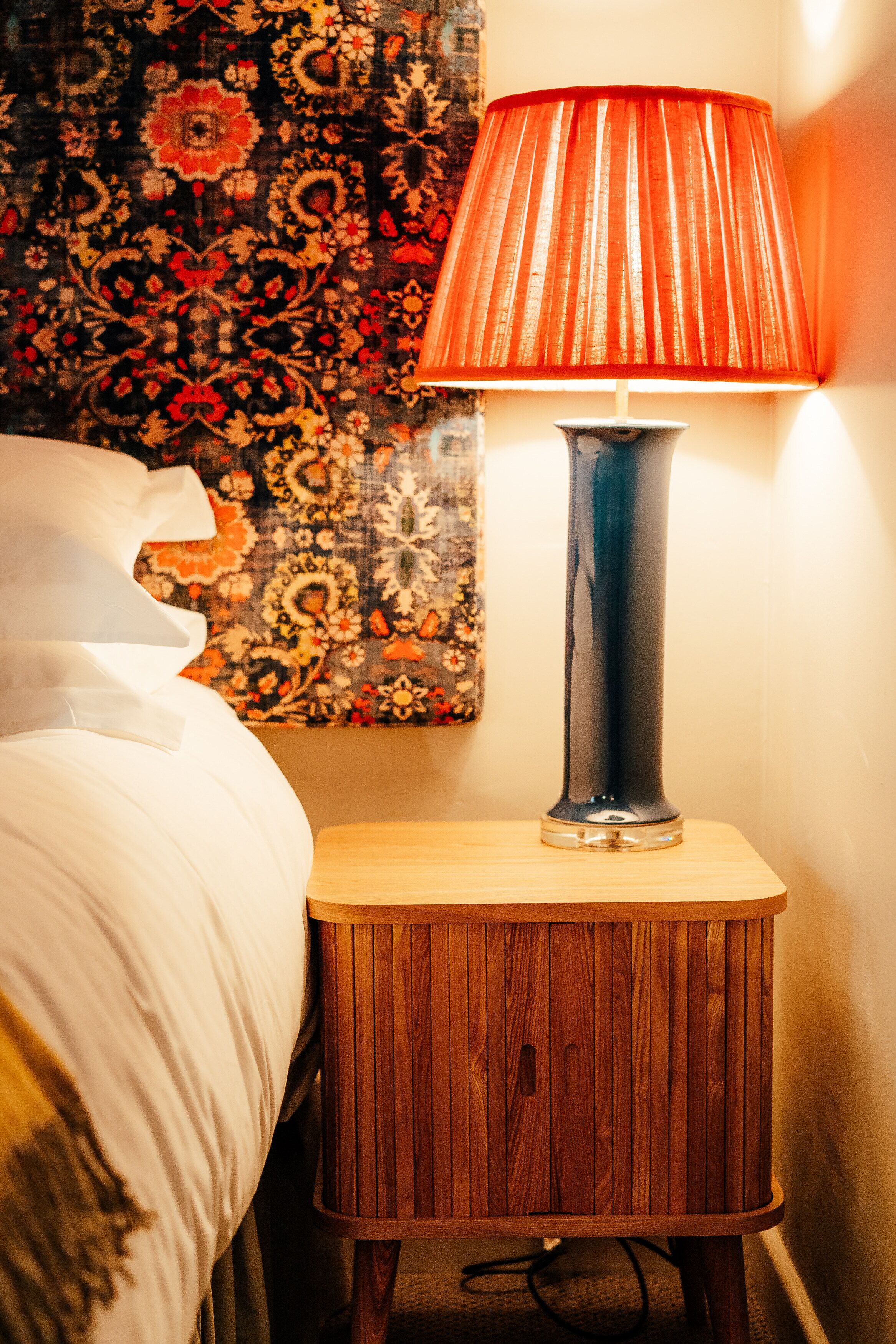 Una cama con ropa blanca, una mesita de noche de madera con una lámpara, y un tapiz floral en la pared.