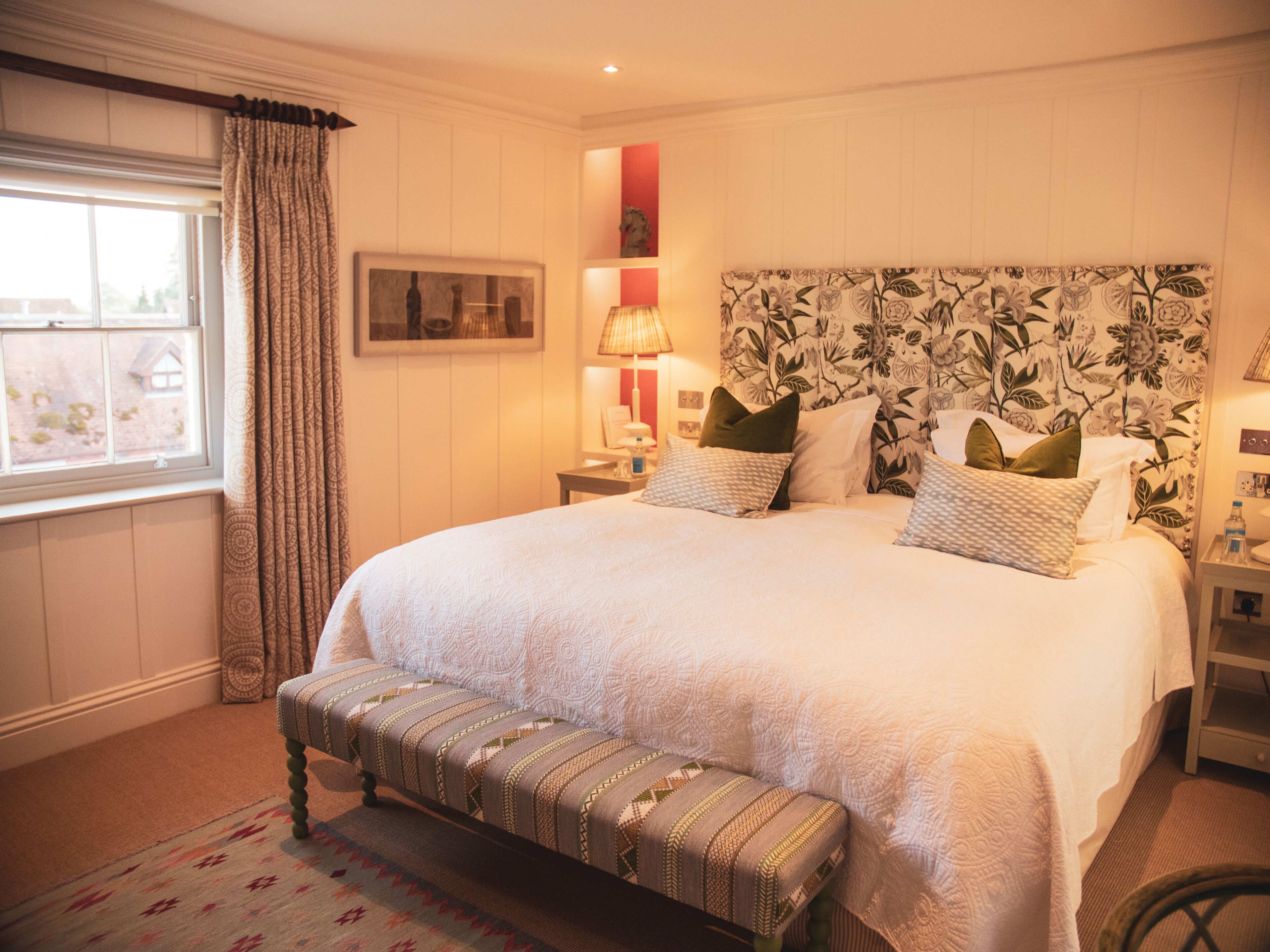 A neatly arranged bedroom with a large bed, a bench, a bedside table, and a window with curtains.