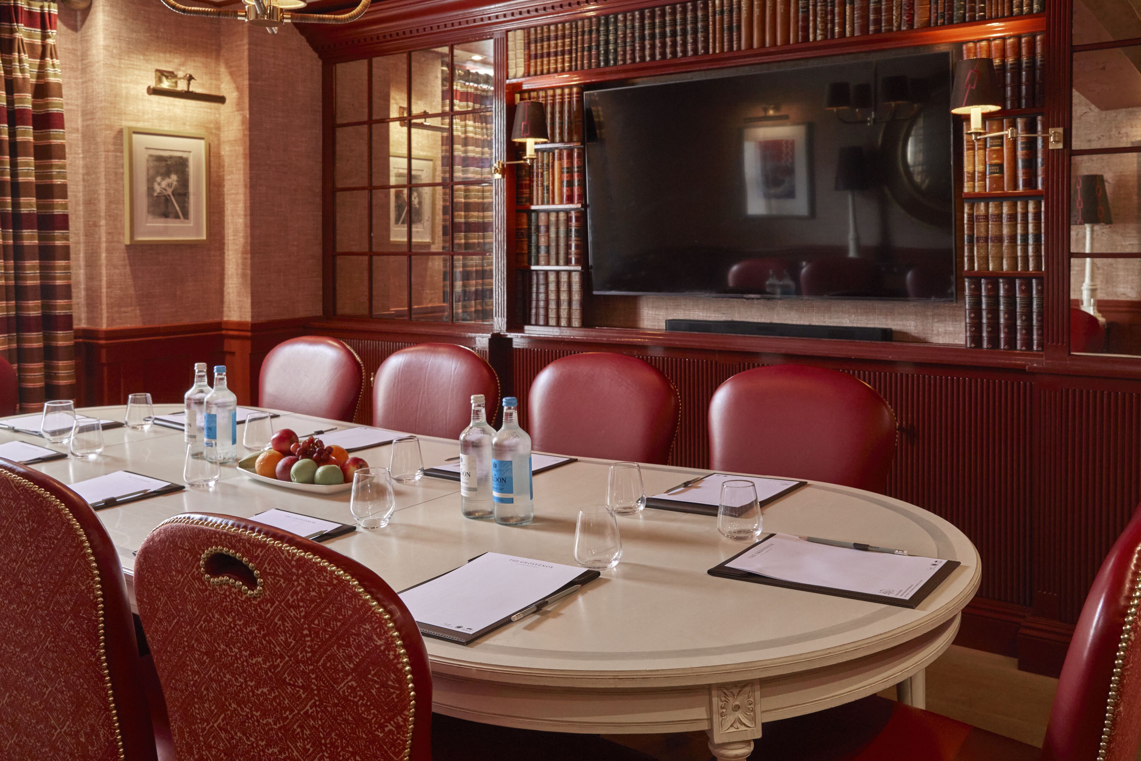 A formal meeting room with a round table, leather chairs, a large flat-screen TV, and bookshelves.