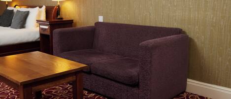 Una habitación de hotel con un sofá morado, una mesa de madera, una alfombra estampada y una cama con ropa blanca.