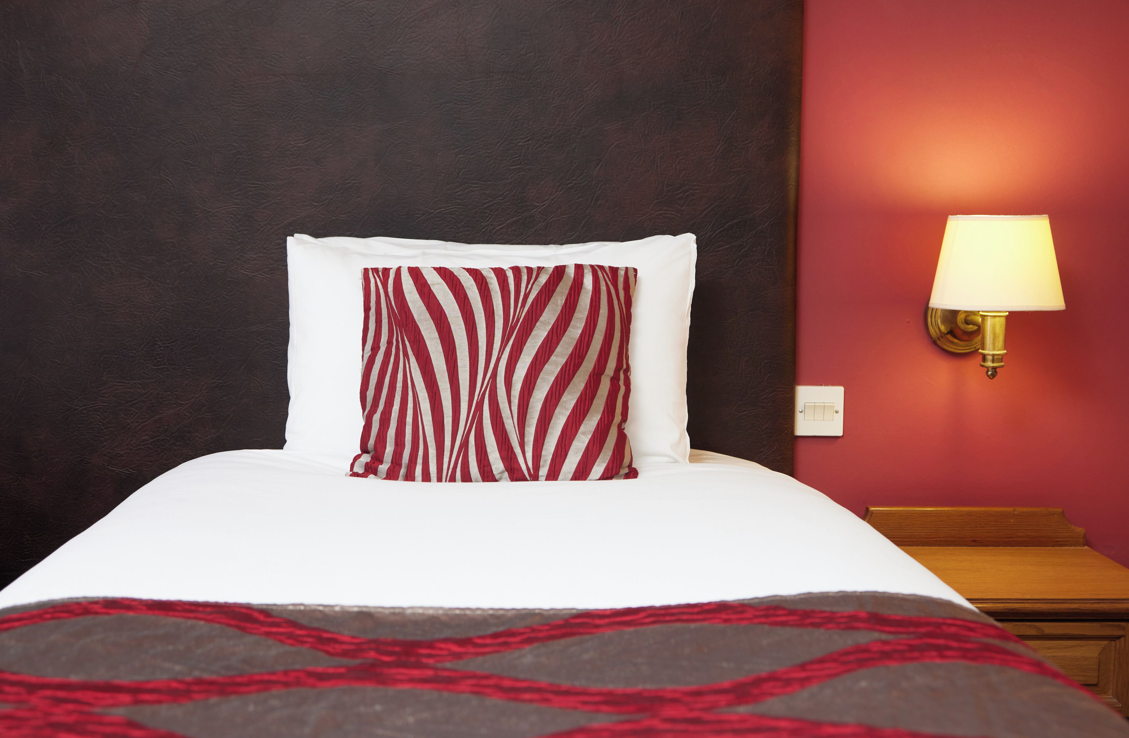 A neatly made bed with a white and red striped pillow, a red and gray blanket, and a wall-mounted lamp on a wooden nightstand.