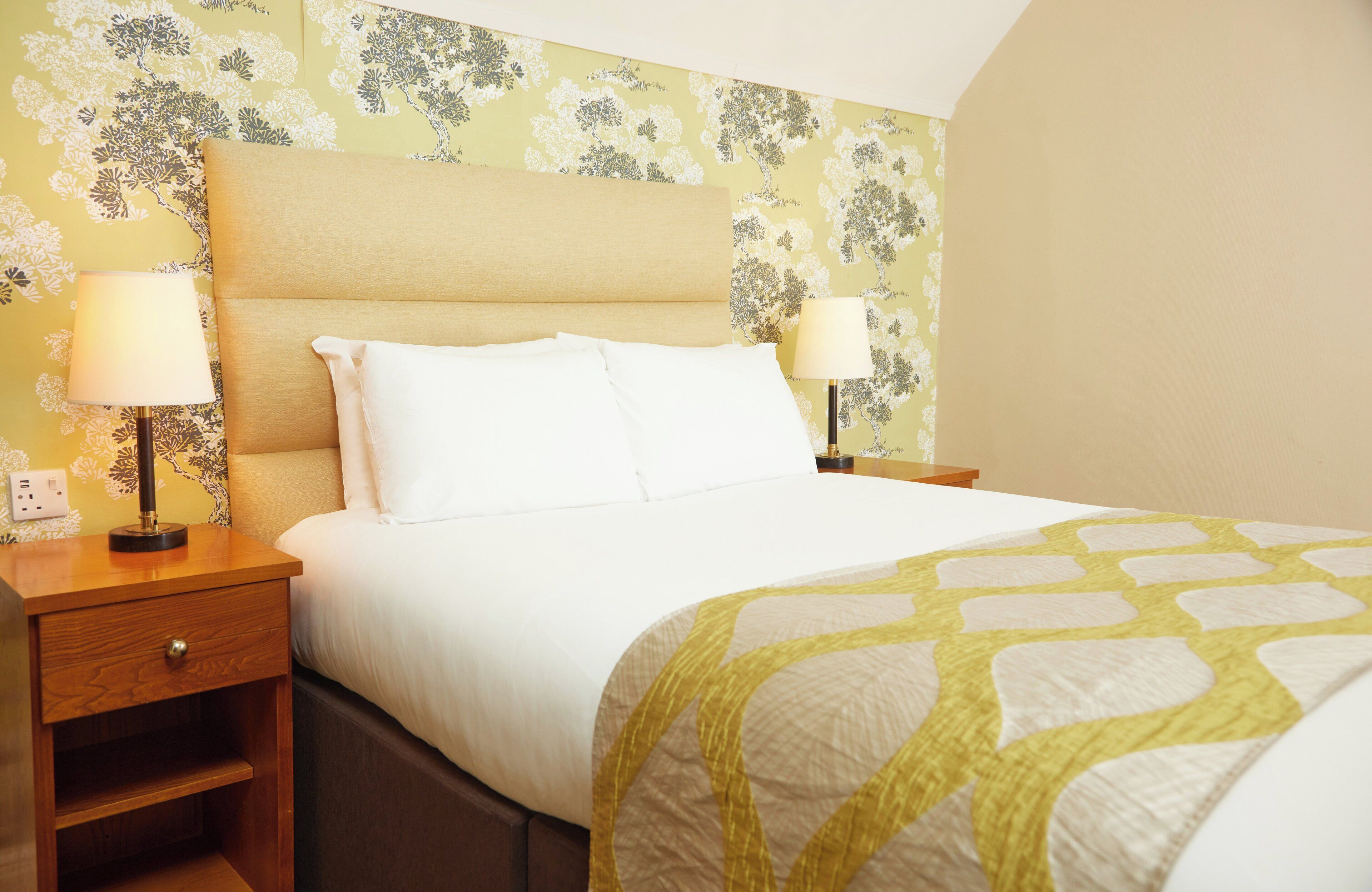 Una cama impecablemente arreglada con ropa de cama blanca y un plaid amarillo estampado, flanqueada por dos mesitas de noche con lámparas, sobre un fondo de papel tapiz con motivos florales.