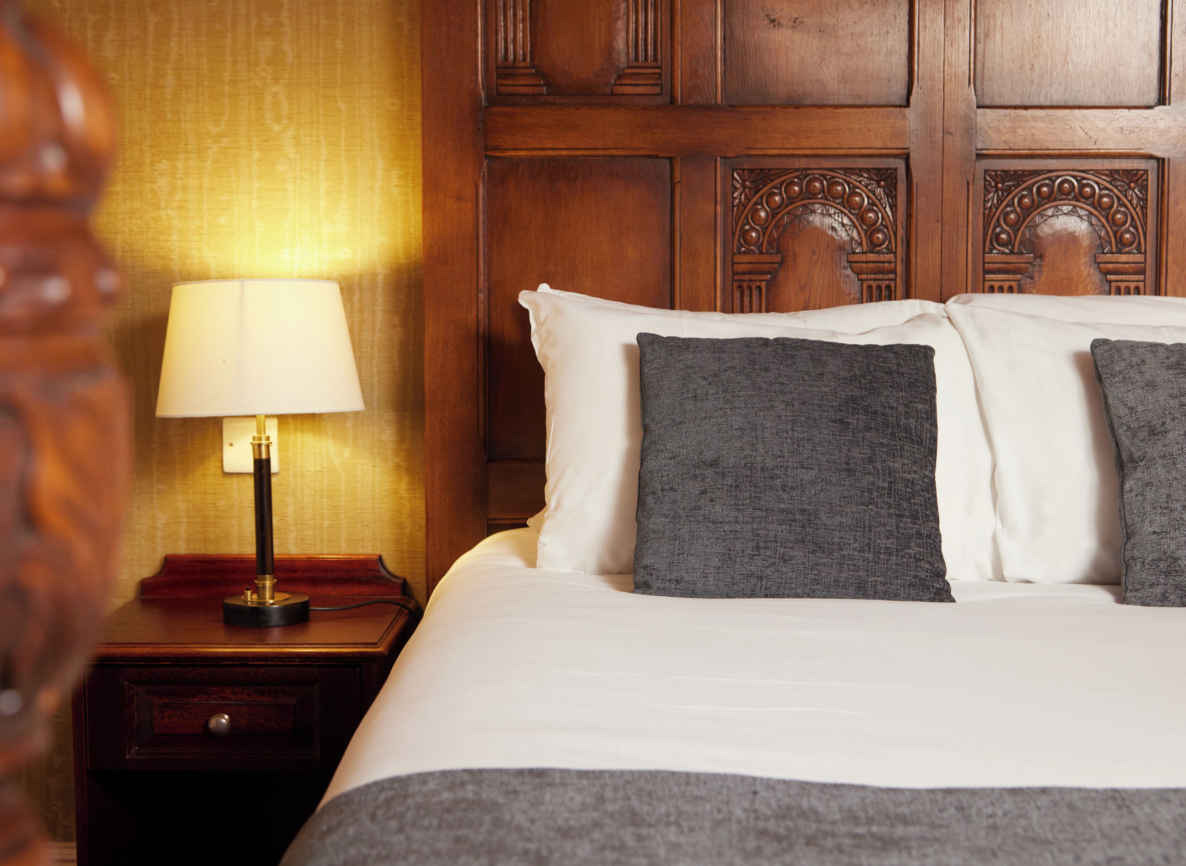 Habitación de hotel con cabecera de madera, lámpara de noche, cama blanca con almohadas grises y mesita de noche de madera con un cajón.
