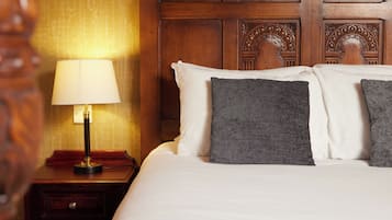 Habitación de hotel con cabecera de madera, lámpara de noche, cama blanca con almohadas grises y mesita de noche de madera con un cajón.