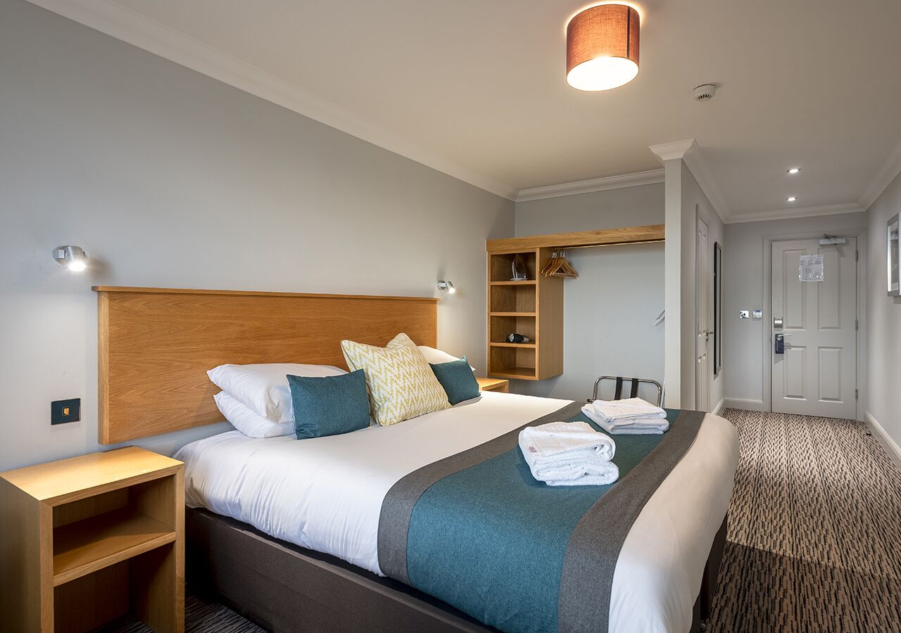 A hotel room with a large bed, a wooden headboard, a bedside table, a wardrobe, and a bathroom door.