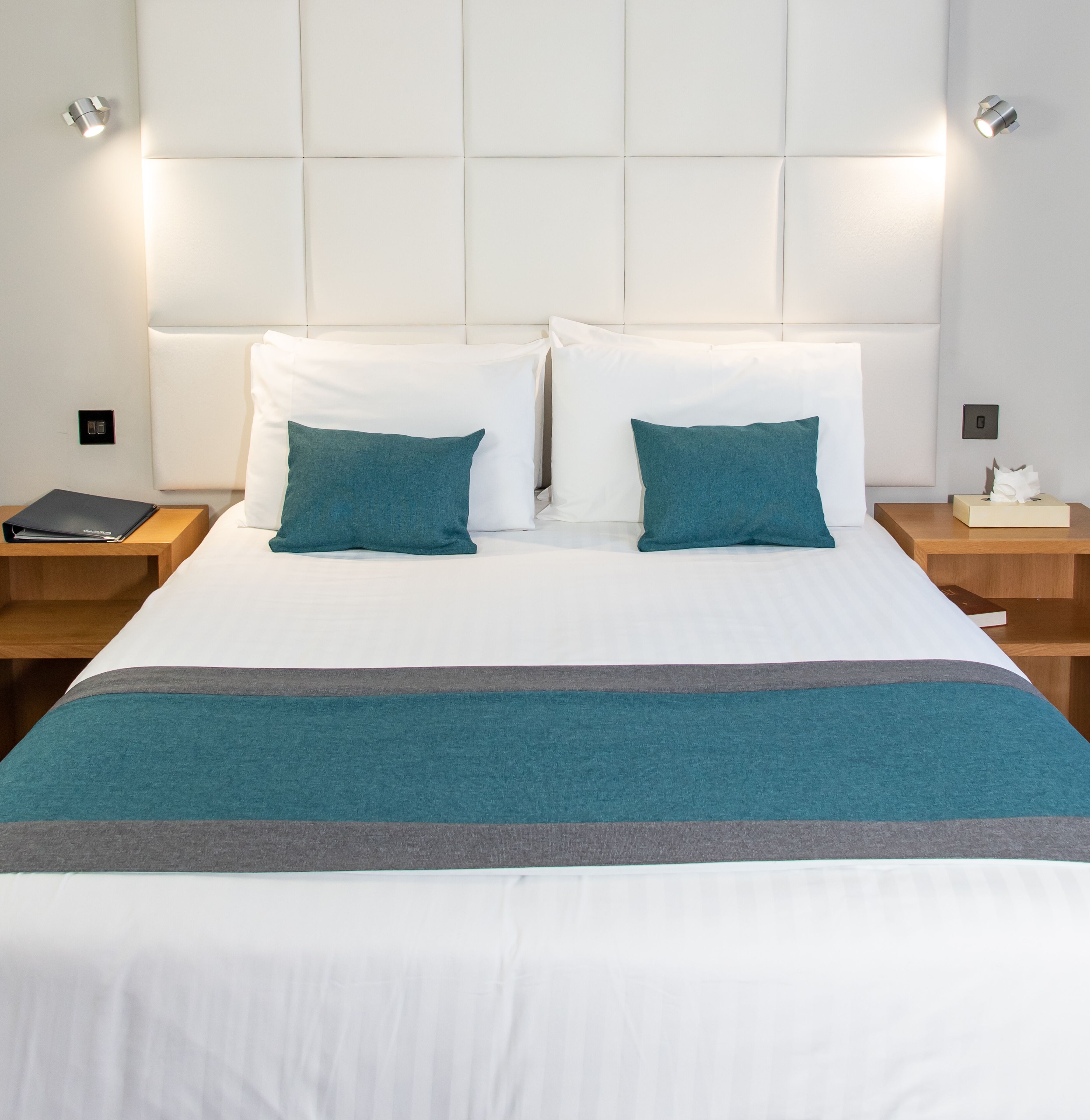 Een net opgemaakt bed met wit beddengoed en teal kussens, een teal- en grijze gestreepte deken, en houten nachtkastjes met een boek en een zakdoos erop.