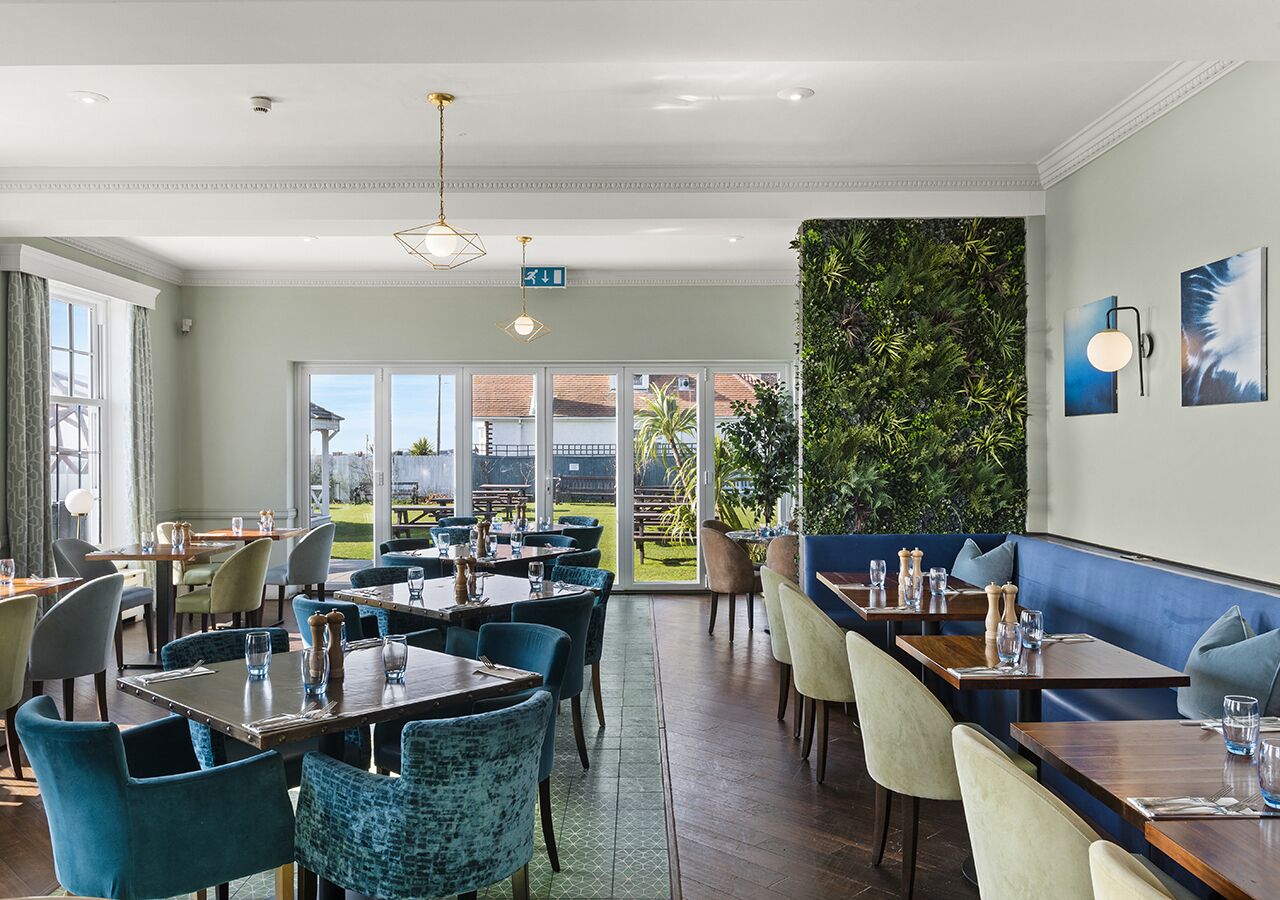 A restaurant with multiple dining tables and chairs, a green wall with plants, and large windows offering a view of the outdoors.