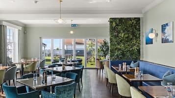 A restaurant with multiple dining tables and chairs, a green wall with plants, and large windows offering a view of the outdoors.