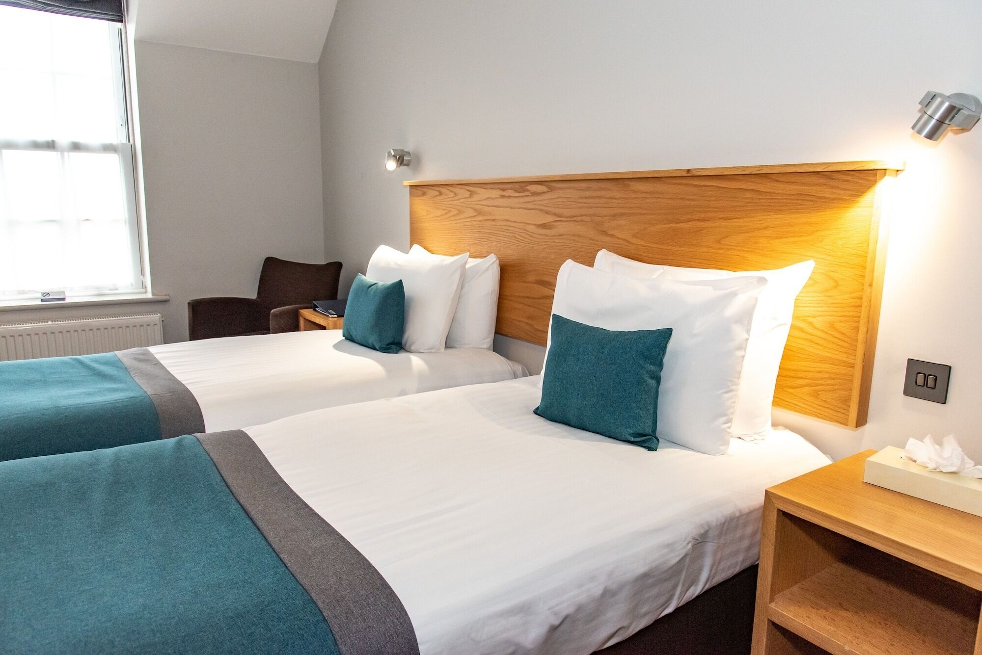 Quarto de hotel com duas camas, cada uma com roupa de cama branca e almofadas verde-água. Possui cabeceira de madeira, mesa de cabeceira com abajur e janela com persianas.